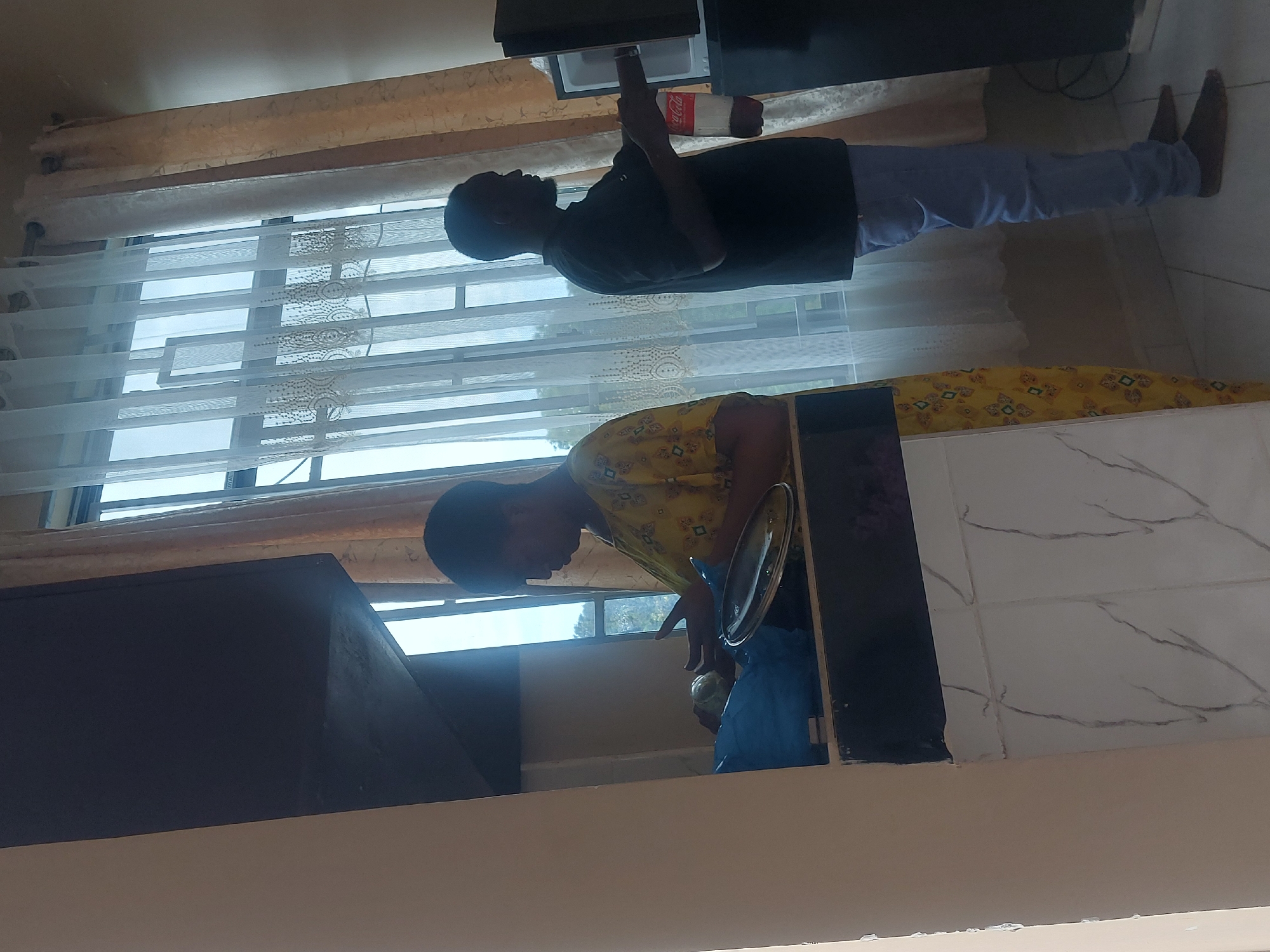 A domestic interior scene is depicted in the image. Two individuals are visible. One is standing and reaching into a cabinet, while holding a bottle. The other person is at a sink, with what appears to be a covered dish. The location cannot be determined from the image.