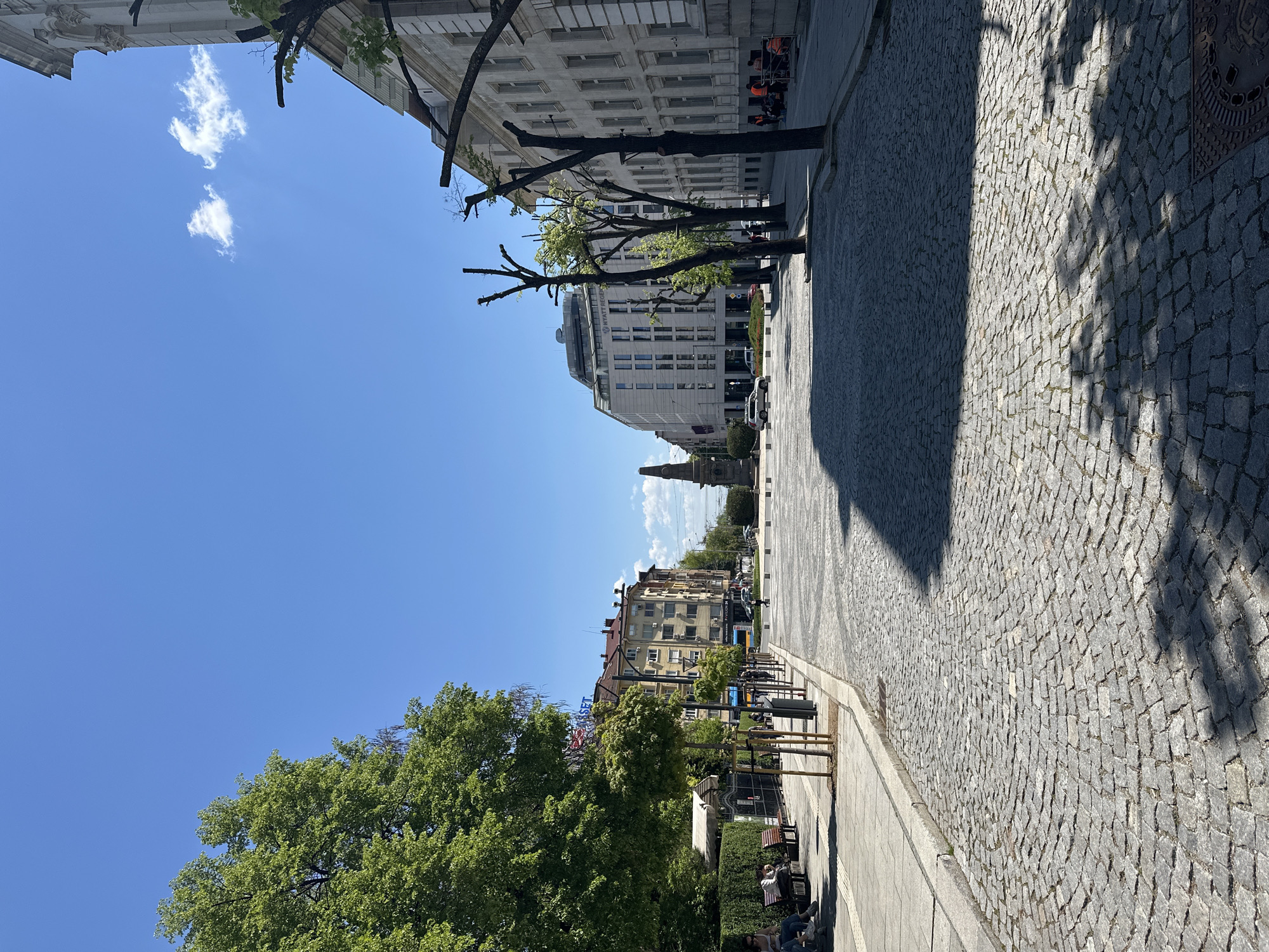 The image depicts a street scene in Sofia, Bulgaria, taken on a sunny day. The view looks down Vitosha Boulevard, a pedestrian street paved with cobblestones. The sky is blue with a few scattered clouds.
Buildings line both sides of the street, ranging in height and architectural style, from older buildings with ornate facades to more modern structures. Trees, some with pruned branches, are planted along the street. Shadows from the trees and buildings stretch across the cobblestone pavement.
Toward the end of the street, the Sveti Georgi Monument is visible.
On the right side of the street, a sidewalk features benches and landscaping. Several people are seated on the benches.