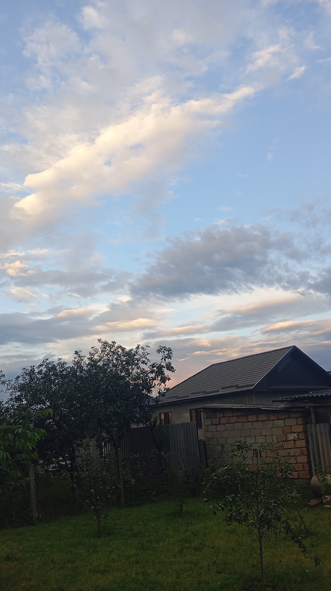 The image shows a backyard scene in Baksan, Russia, under a cloudy sky at dusk. The foreground is dominated by a lush green lawn with several small trees and bushes. A large, mature tree stands to the left, its dark foliage silhouetted against the sky. Behind the trees, a corrugated metal fence runs across the middle ground, separating the yard from the structures beyond.
To the right of the fence, a building with a dark, gabled roof is visible. Part of the building is constructed with light-colored bricks. The overall impression is one of a rural or suburban residential setting.
The sky is a prominent feature, filled with a mix of light and dark clouds. Some clouds are illuminated by the setting sun, casting a warm, golden glow, while others appear darker and more dramatic. The soft lighting and cloud formations suggest the late evening hours. There is no text visible in the image. The scene evokes a sense of quietude and the natural beauty of the transition from day to night.