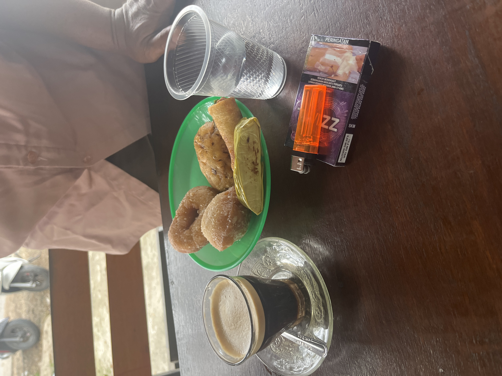 In Keudematangkuli, Indonesia, a dark brown wooden table is the setting for a casual outdoor scene. A clear glass of dark coffee with a thick crema sits on an ornate glass saucer with a spoon. Next to the coffee, a green plastic plate holds several sweet snacks, including two ring-shaped donuts coated in sugar and two fried pastries, possibly pisang goreng or similar fried bananas. Adjacent to the plate is a clear plastic cup filled with water. In the foreground, a pack of cigarettes, identified by the "ZZ" logo and Indonesian warning labels, lies next to a bright orange lighter. Partially visible in the background is a person wearing a pink shirt, and beyond that, two motorcycles are parked.