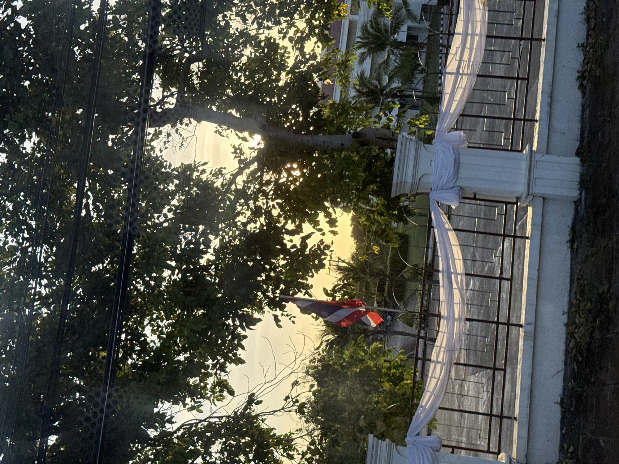 The image depicts an outdoor scene at dusk, likely in Baan Tha Chat Chai, Thailand. A large tree with lush green leaves dominates the upper portion of the frame, with power lines running horizontally through the branches. The setting sun casts a warm, golden glow through the foliage, creating a silhouette effect. In the mid-ground, a Thai flag is visible, mounted on a pole, with its red, white, and blue stripes distinct against the bright sky. A decorative white fabric drapes over a metal fence, which is supported by white pillars. Beyond the fence, palm trees and other greenery are visible, along with a white building with a red-tiled roof. The foreground shows a dark, unpaved area with scattered leaves. The overall impression is a tranquil, natural setting illuminated by the fading sunlight.