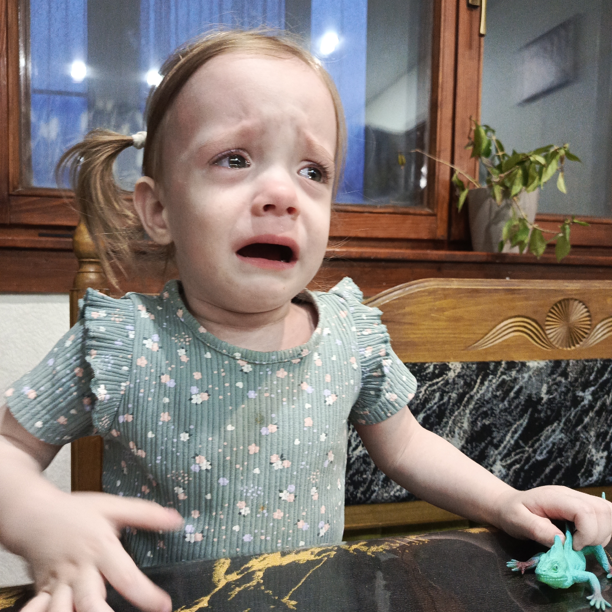 A young Caucasian girl with blonde hair styled in pigtails is crying, tears visible on her face. She is seated at a table with a dark, marbled pattern and gold accents. She wears a light green, floral-patterned shirt with ruffled sleeves. A green toy lizard is on the table next to her right hand. In the background, there is a window with a wooden frame and a potted plant on the windowsill. The location is indoors, but there is no indication it is in Марковский, Russia.