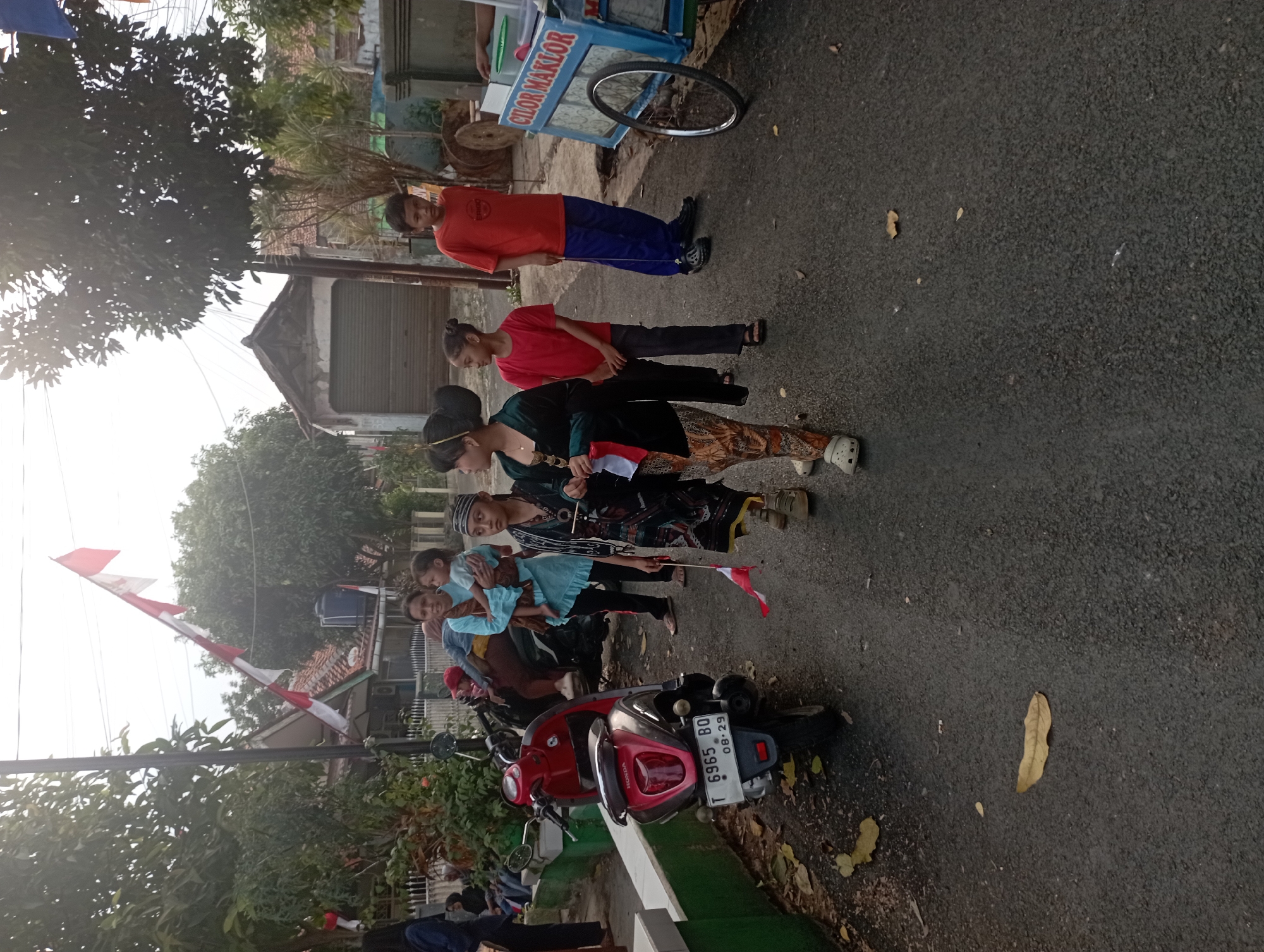 In an outdoor scene, possibly in a residential area, several individuals are present. A woman in a light blue top is holding a young child. Two girls, one in a green jacket and patterned pants, and another in a red t-shirt and grey pants, are standing near each other. A boy in an orange t-shirt and blue pants is also visible. A red scooter is parked on the left side of the frame, with a license plate reading "T 6965 BQ 08-29". In the background, a cart with "CILOR MAKLOR" written on it is present, along with a bicycle wheel. Indonesian flags are displayed, some draped from a pole and others held by individuals, suggesting a festive or nationalistic occasion. The ground is paved asphalt, with some fallen leaves. The environment appears to be a street with houses and trees.