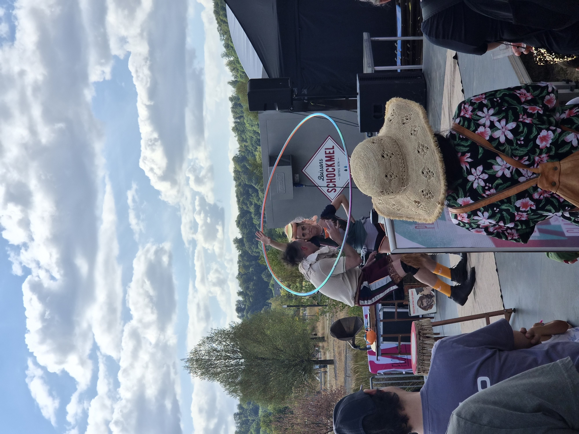 This image captures an outdoor performance scene in Sanem, Luxembourg. In the foreground, the backs of several spectators are visible, with one person in a wide-brimmed straw hat and floral shirt facing away from the camera. The central focus is a performance taking place on a low stage. A male performer, wearing a light-colored shirt, dark shorts with patterned trim, and bright orange socks, is actively engaged with a female performer. The female performer, with pink hair and wearing athletic shorts and a top, is holding a colorful hula hoop that encircles them. Behind the performers, a grey structure displays a diamond-shaped logo for "Boissons SCHOCKMEL - DEPUIS 1974". Speakers and a tent are also present on the stage area. The background reveals a wooded hillside under a partly cloudy sky, suggesting a natural or park-like setting. The overall atmosphere appears to be a casual outdoor event.