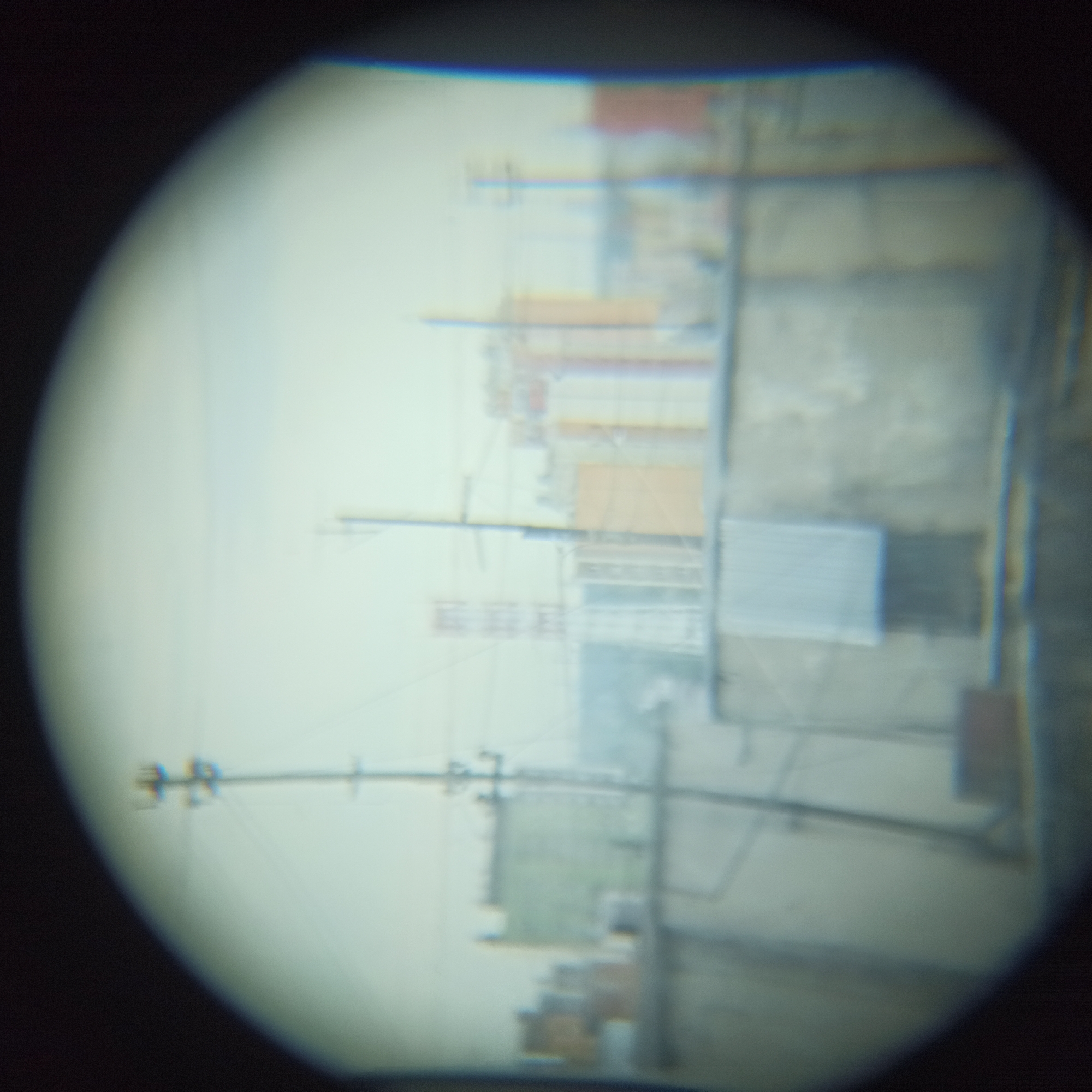 The image, viewed through a circular lens, shows a blurred cityscape with multiple buildings and utility poles. In the foreground, a gray, textured wall runs horizontally across the frame. Above this wall, various structures are visible, including a light-colored building with a distinctive horizontal band and a rectangular object that appears to be a vent or air conditioner unit. To the left, several brick-colored structures are dimly discernible.
Further back, a tall utility pole with horizontal crossbars stands prominently. Behind it, a series of buildings with distinct vertical lines, possibly indicating floors or windows, are visible. One building in the middle ground appears to be light orange or tan. The sky is overcast and pale. The overall impression is of an urban environment, potentially an industrial or residential area, viewed from a distance or through a device with low optical clarity. Without specific identifying landmarks or textual information within the image, it is not possible to definitively determine its location within Nizhny Novgorod, Russia.