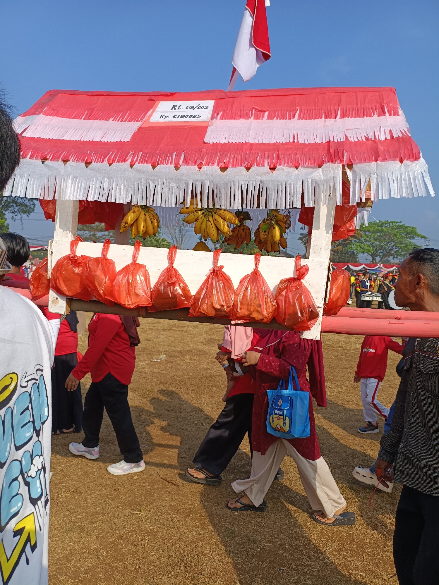 An outdoor daytime scene in Cibodas, Indonesia, depicts a procession. A mobile vendor stall, adorned with red and white striped fabric streamers and a red and white flag, is being carried on poles by several individuals. The stall displays bunches of bananas and several red plastic bags containing unseen items. In the background, a group of people in red uniforms with musical instruments, possibly a marching band, are visible. The ground is covered in dry grass, and the sky is clear blue. The people in the foreground are dressed in casual attire, including a person in a white shirt with colorful graphics and another in a dark striped shirt.
