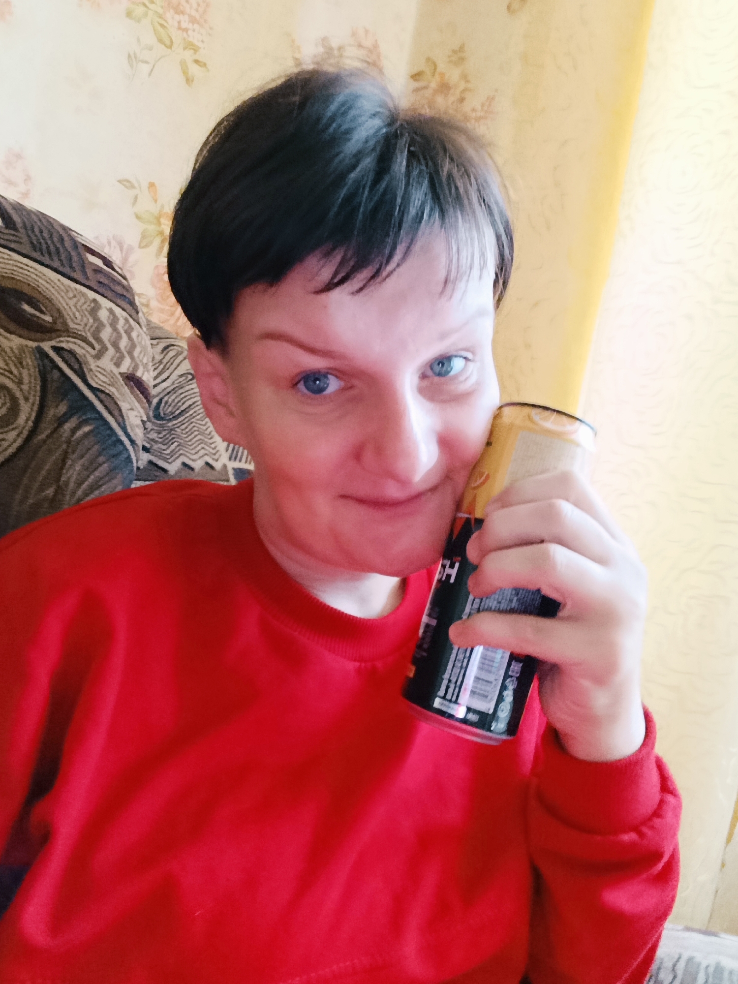 A medium shot shows a person with short, dark hair and blue eyes holding a can of what appears to be an energy drink to their right cheek. The person is wearing a red sweatshirt. The background consists of a wall with floral wallpaper and a yellow curtain. A portion of a patterned couch is visible on the left side of the frame. The scene is indoors, likely in a residential setting. There is no information available about the location being in the city of Фоки, Russia.
