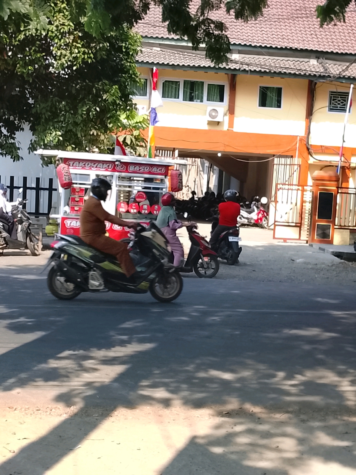 The image shows a street scene in Slawi, Indonesia, on a sunny day. A food cart with the words "Takoyaki & Baso Aci" is visible on the left side of the image. Several people are on motorcycles. One person in a brown outfit and helmet is riding a green and black motorcycle. Another person in a light blue outfit and pink headscarf is on a red and black motorcycle. Further in the background, there are more motorcycles parked, and a building with yellow and orange accents. The scene is illuminated by sunlight, casting shadows from trees onto the asphalt road.