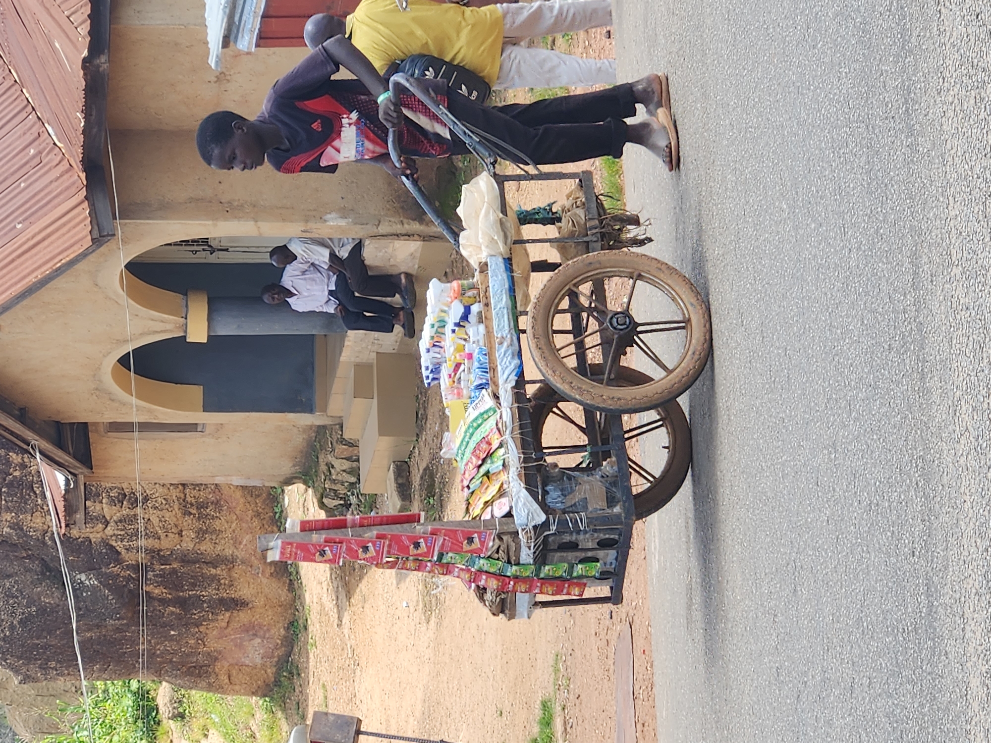 In Kabong, Nigeria, a young Black male, wearing a black and red t-shirt and black pants, pushes a two-wheeled cart laden with various goods, including packaged snacks and drinks. The cart has a metal frame and wooden shelves. The boy is wearing sandals. In the background, two Black males, dressed in light-colored shirts and dark pants, are seated on a step in front of a building with a corrugated metal roof and arched doorways. Another Black male, wearing a yellow shirt and white pants, stands to the right. The setting appears to be a street with a paved road and a dirt area with vegetation and a large rock formation.