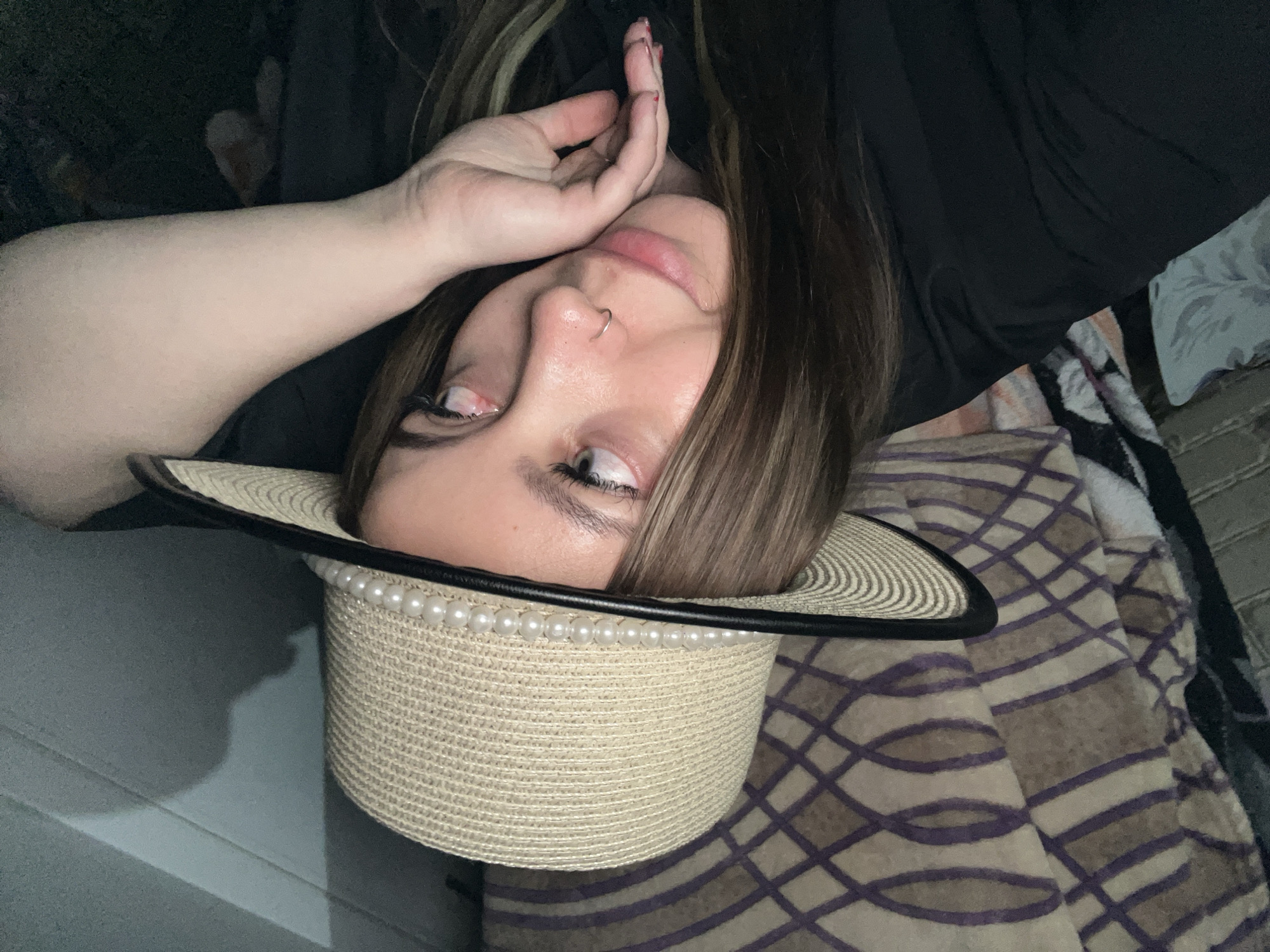 The image shows a woman lying down, likely on a bed or couch, wearing a hat with a black brim and pearl embellishments. She is wearing a black top and has her hand near her face. The background includes a patterned blanket. No specific location within Novopokrovskaya, Russia, can be determined from the image.