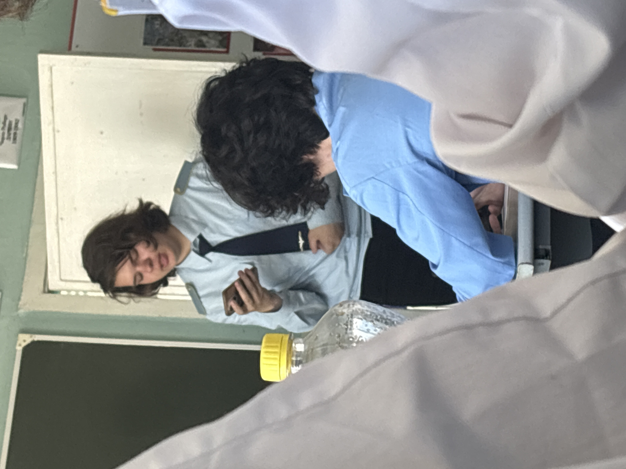 The image captures an indoor scene, appearing to be a classroom, viewed from a slightly elevated and obscured perspective. The original image is rotated 90 degrees counter-clockwise.
In the mid-ground, a person with dark, curly hair is seen from behind, wearing a light blue long-sleeved shirt. Their body is angled forward and to the left. Their hands are partially visible near their lap.
To their left, another individual is visible, looking towards the camera with a neutral to slightly amused expression. This person has medium-length brown hair and is wearing a light gray long-sleeved uniform shirt with a dark tie and visible shoulder epaulets or straps. They are holding a dark-colored smartphone in their hands.
Between these two individuals, on a surface like a desk, a clear plastic bottle with a bright yellow cap is present.
The background consists of a light-colored board or whiteboard above a dark green or black chalkboard. A small white paper with very faint, illegible text is affixed to the wall on the far left.
The foreground is partially obscured by a large area of light-colored fabric, likely the clothing or sleeve of someone sitting in front of the camera, suggesting a candid photo taken within a school or similar educational setting. No discernible text is clearly visible on any of the boards or papers.