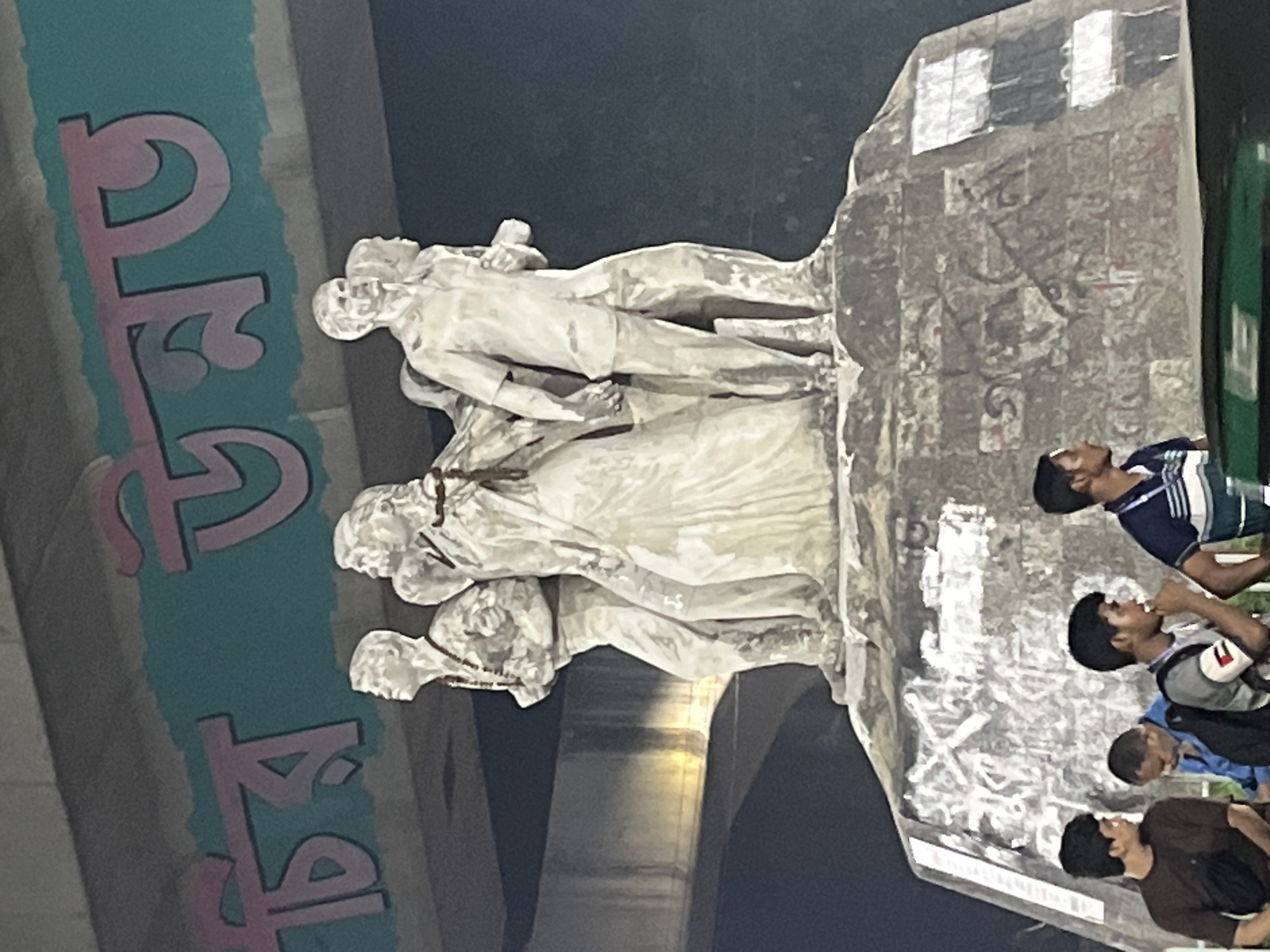 The image shows the Aparajeyo Bangla sculpture at the University of Dhaka in Bangladesh. The sculpture depicts three students, a man and two women, representing the struggle for independence. In the foreground, several young men appear to be watching or looking up at the sculpture, some possibly eating. Graffiti in Bengali script is visible on a structure to the left of the sculpture. The scene appears to be under an overpass or bridge, with architectural support structures visible in the background.