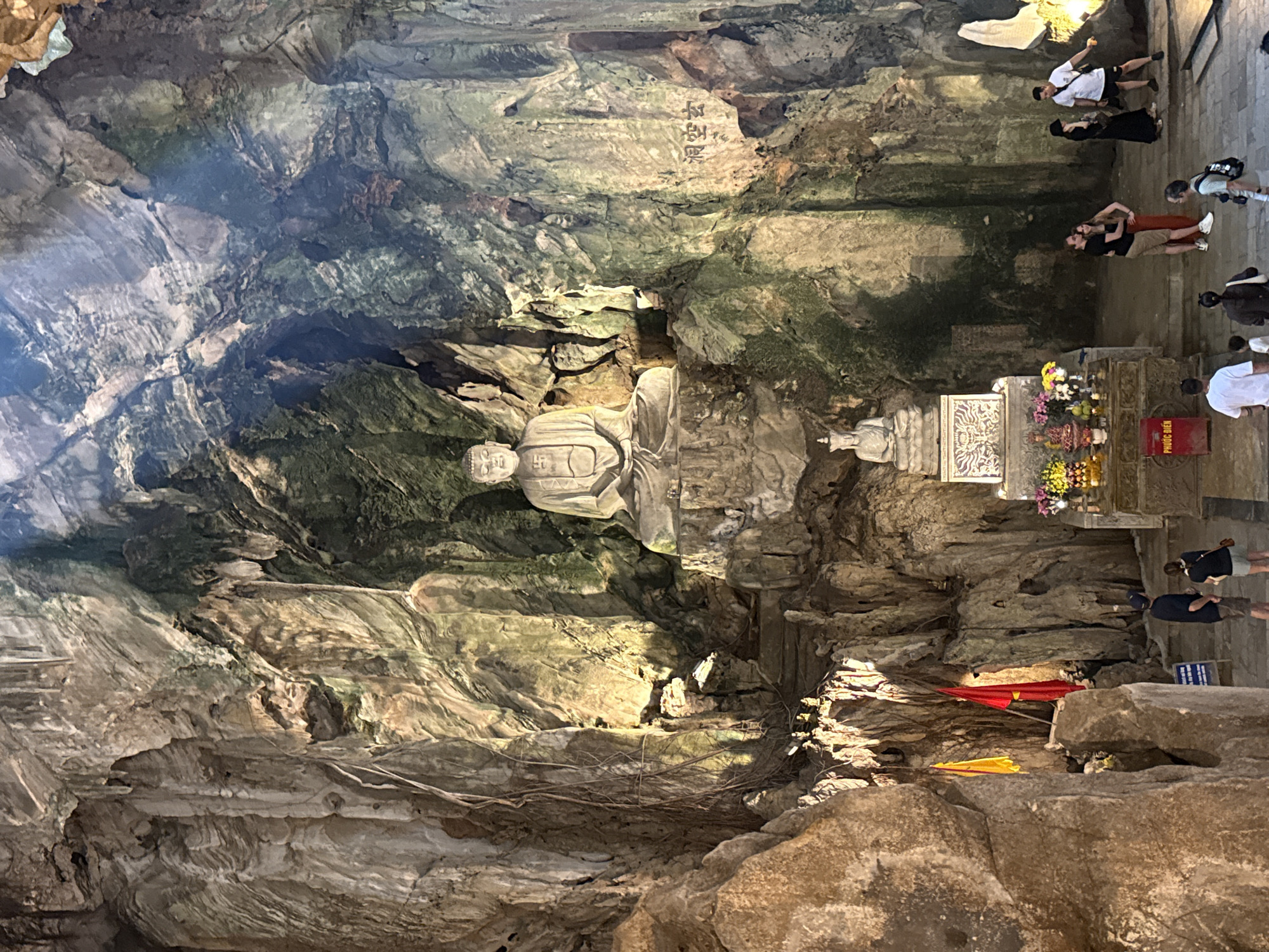 This image depicts the interior of Huyền Không Cave (Huyền Không Động), located within the Marble Mountains (Ngũ Hành Sơn) in Đà Nẵng, Vietnam. The scene is a large, natural limestone cave, characterized by rugged, uneven rock formations, stalactites, and visible root systems clinging to the walls. Natural light filters in from a large opening in the cave ceiling, supplemented by artificial spotlights illuminating key features.
In the upper-center, a large, serene, seated Buddha statue carved from light-colored stone is positioned high on a natural rock platform. The Buddha features a swastika symbol on its chest. Below and slightly to the left of this main statue, a smaller, similarly styled seated Buddha rests on an ornate, tiered altar. This altar is adorned with various offerings, including colorful fresh flowers (pink, purple, yellow), fruits, and other devotional items. A red donation box, labeled "PHƯỚC ĐIỀN," is visible on the floor in front of the altar. To the right of the main Buddha, Chinese characters (洞空玄 - Huyền Không Động) are carved into the cave wall.
On the left side of the cave floor, near a rock formation, stand two flags: one yellow and one red. A series of stone steps ascends the rocky terrain towards the platform of the large Buddha.
Approximately ten to twelve individuals, appearing to be tourists or visitors, are present on the paved cave floor. Most are casually dressed. A couple is embracing near the center-right of the frame. Other individuals are observed standing, walking, or looking around. One man in a white t-shirt and shorts points towards an area off-camera to the right. The overall environment is dimly lit, with illuminated sections highlighting the religious statues and rock formations, creating a contemplative atmosphere within the natural cavern.