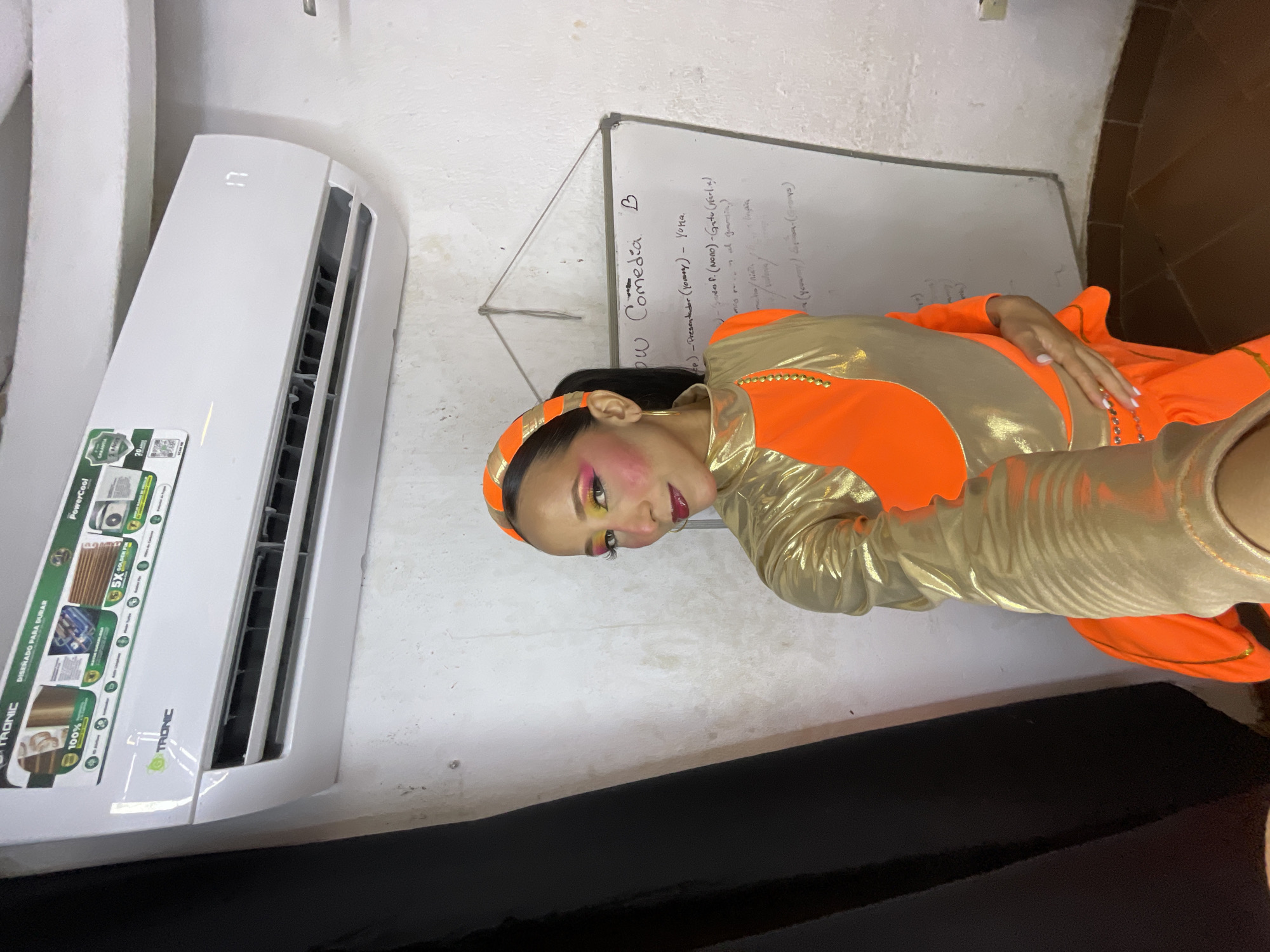 In a room with white walls and brown tiled flooring, a woman with dark hair, wearing an orange headband, bright pink and yellow eye makeup, and a shiny gold and orange costume, stands facing the camera. She is holding a phone in her right hand, presumably to take a selfie. To the left, a black curtain hangs. Above her, a white air conditioner unit with a digital display showing "17" is mounted on the wall. To her right, a whiteboard displays handwritten text in black marker, including "SHOW Comedia B". The overall scene suggests a backstage or preparation area, possibly for a performance.