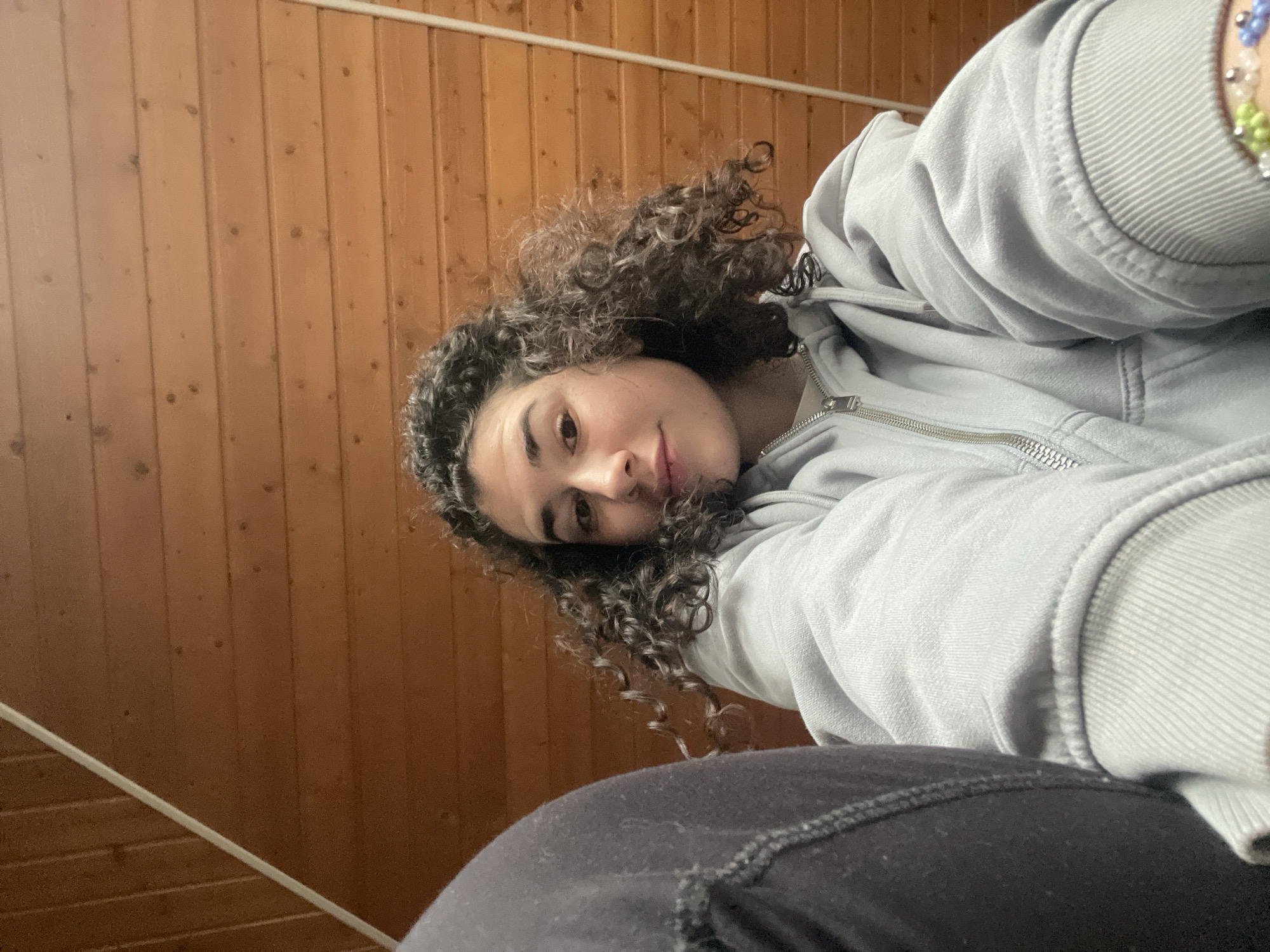 A young person, appearing to be female, is captured in a close-up, low-angle self-portrait indoors. She has medium-toned skin and abundant dark, curly hair that frames her face. Her gaze is directed straight at the camera, and she has a calm, gentle expression with a hint of a smile.
She is dressed in a light grey hooded sweatshirt with a zipper down the front and appears to be wearing dark-colored pants or leggings. On her right wrist, a multi-colored beaded bracelet is visible, featuring shades of blue, green, and white.
The background consists of warm, light brown wooden paneling, possibly a wall or a sloped ceiling, indicating an indoor setting. A thin, light-colored horizontal rod or pipe runs across the upper part of the wooden background. The overall scene suggests a casual moment, perhaps relaxing in a room in Sofia, Bulgaria, given the context. There is no visible text in the image.