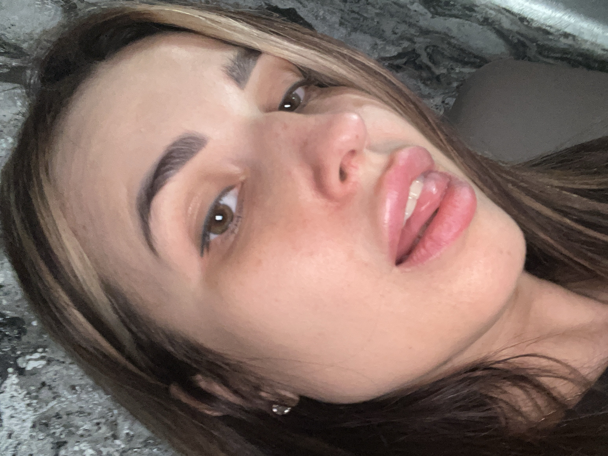 A close-up, tilted portrait depicts a woman with light brown hair and brown eyes. Her complexion is fair, and her lips are full and pink. She appears to have well-defined eyebrows. An earring is visible in her ear. The background is a grey and white textured surface, possibly a countertop or similar.