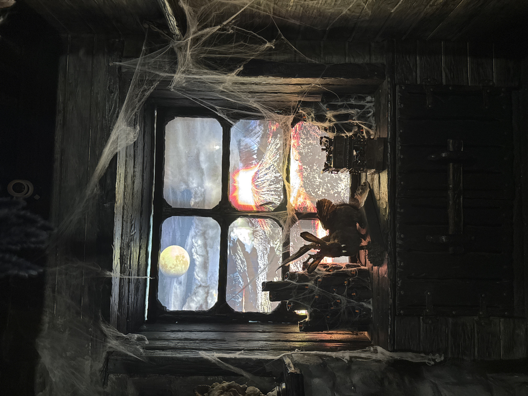 The image depicts a dark, aged wooden window frame covered in artificial cobwebs. Through the panes, a backdrop is visible: one pane shows a moonlit scene, while the others display fiery, abstract imagery suggesting a Halloween or horror-themed display. A large, artificial spider sits on a platform within the window. The setting appears to be indoors. There is no indication of the city of Kazan, Russia.