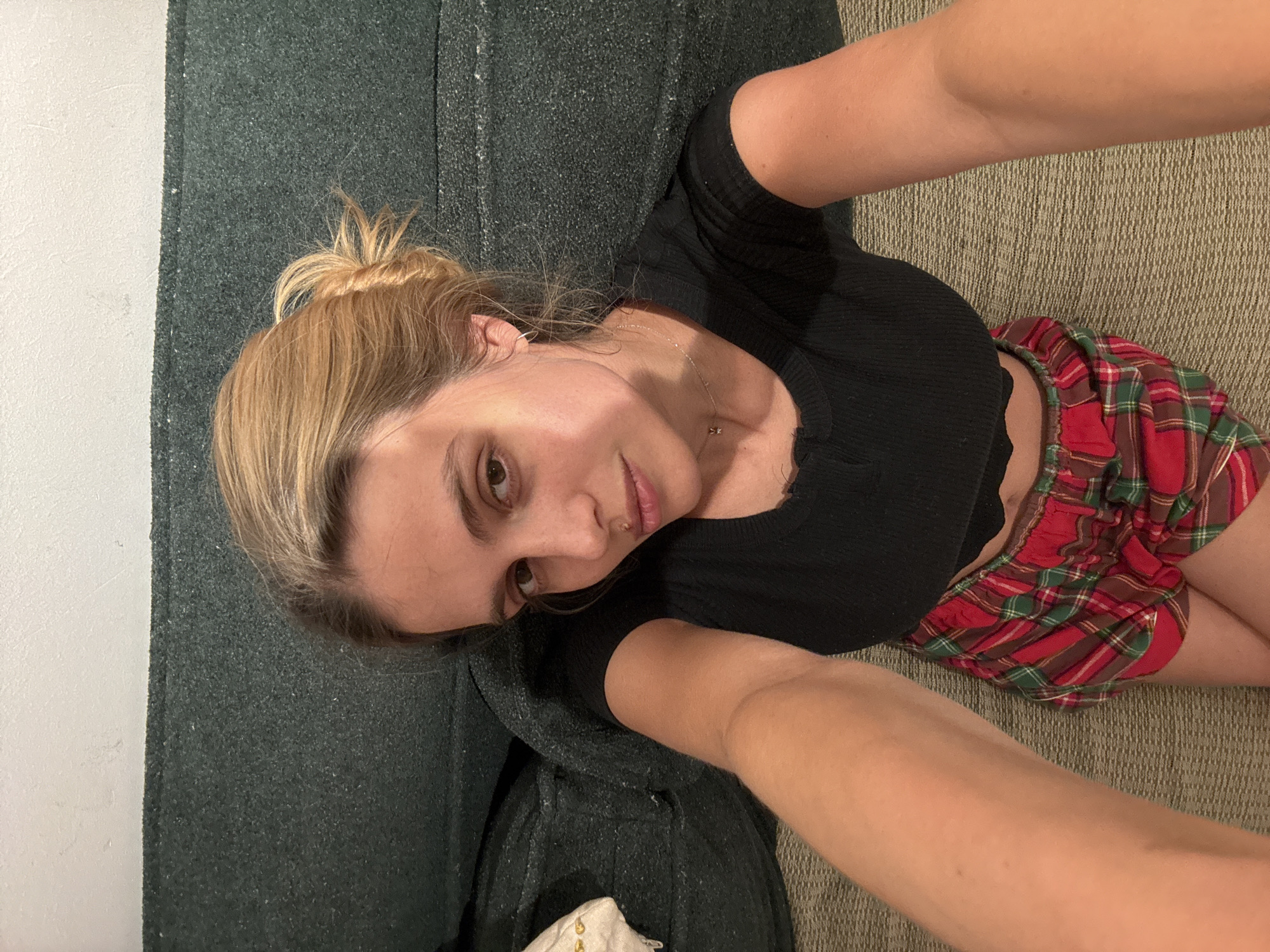 A person is lying on a couch, taking a selfie. The person has blonde hair pulled back in a bun, is wearing a black short-sleeved shirt, and plaid red and green shorts. The couch has a dark green textured fabric on the back and a beige textured fabric on the seat. A white wall is visible in the upper left corner. The location is unknown.