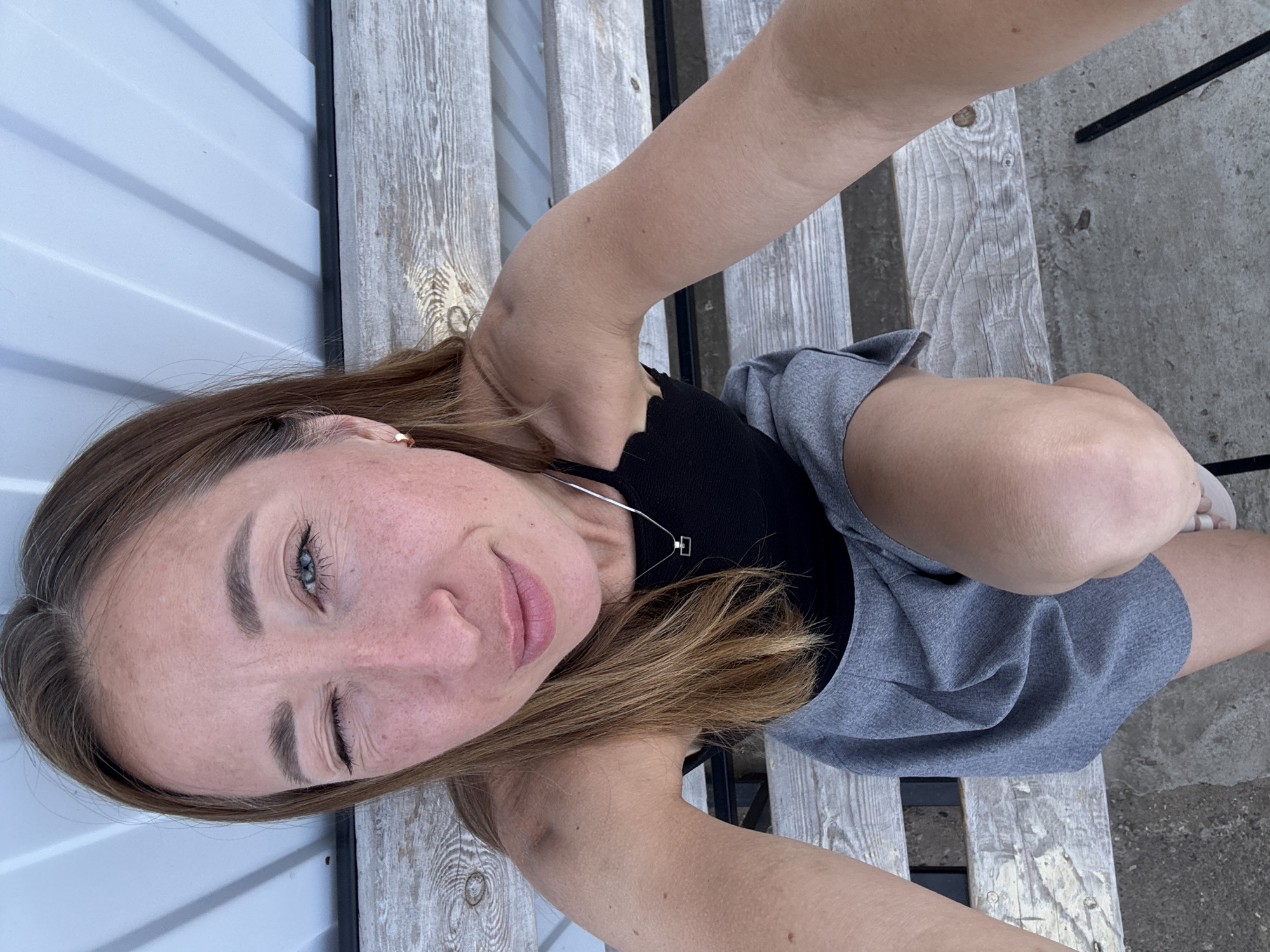 A woman is taking a selfie while sitting on a wooden bench in Chaikovsky, Russia. She has light brown hair and blue eyes, and she is winking at the camera. She is wearing a black top and a gray skirt. There is a white metal structure to her left and a concrete area to her right. The bench appears to be outdoors.
