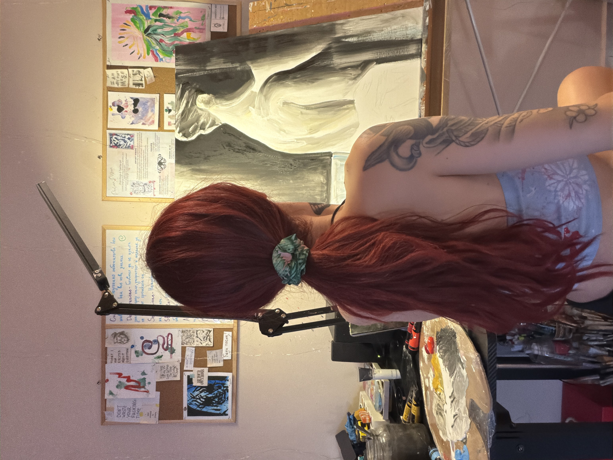 The image depicts a person with long, dark red hair, seen from behind, working in an art studio. The studio appears to be a domestic space, possibly an apartment, with art supplies and personal creative materials displayed on corkboards and a desk. The person is wearing a light blue and white floral-patterned top.
On the corkboards, various papers with handwritten text, drawings, and prints are pinned. Some of these appear to be motivational quotes or artistic inspirations. One paper has the text "DON'T WASTE YOUR FUCKING TIME." Another document, titled "Padlurn Pitan," outlines information about "Paintings & Textile Art," including "ARTIST VISION" and "VALUES."
In front of the person is an easel holding a large, unfinished painting of what appears to be a classical sculpture, rendered in black and white and muted yellow tones. A wooden palette with various colors of paint is visible on a desk to the left, alongside jars, tubes of paint, and brushes. The desk also holds a lamp that illuminates the workspace.
The overall environment suggests a dedicated artist's personal workspace, filled with creative endeavors and inspirational materials. There is no visual information present to indicate the specific location within the city of Svilengrad, Bulgaria.