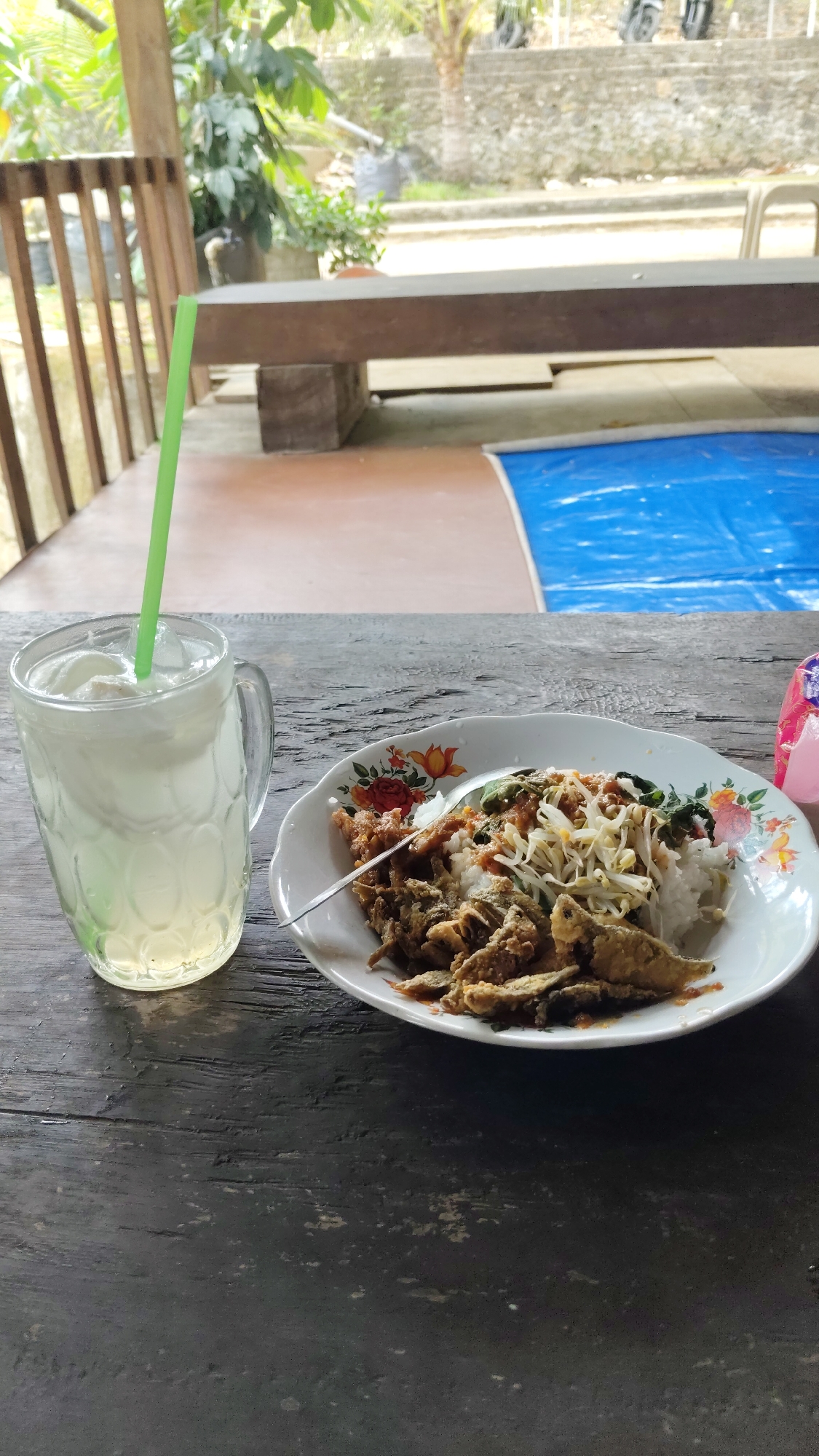 An overhead shot features a meal and drink on a wooden table outdoors. On the left, a tall glass mug is filled with a pale yellow, cloudy beverage and ice cubes, with a bright green plastic straw extending from it. To the right of the mug is a white, floral-patterned plate filled with rice, topped with bean sprouts, leafy greens, and fried fish. A spoon rests on the plate. The table itself is dark-colored and appears weathered. In the background, a wooden bench and a blue tarp are visible, with foliage and a stone wall suggesting an outdoor setting, possibly in Kresek, Indonesia.