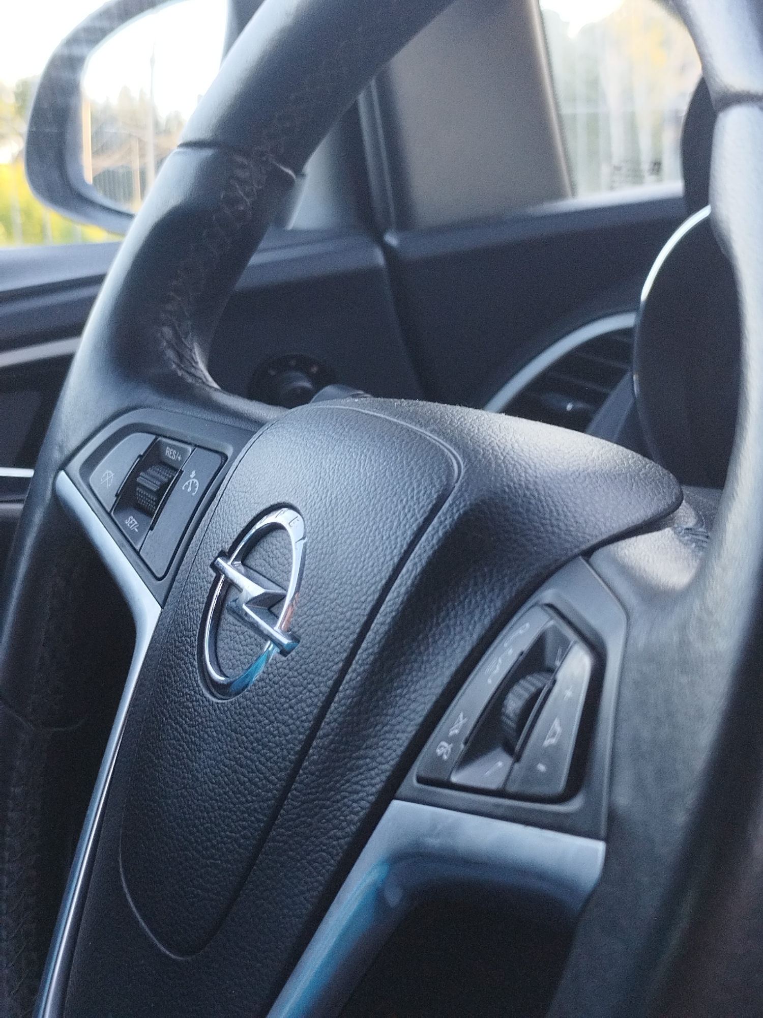 The image shows a close-up of a car's steering wheel. The steering wheel is black and textured, with the Opel logo in the center. There are various buttons on the steering wheel, including controls for cruise control and audio. The interior of the car is visible, with a side mirror and part of the dashboard. The background is blurred, suggesting the car is in motion or parked outdoors. The overall impression is one of a modern car interior.