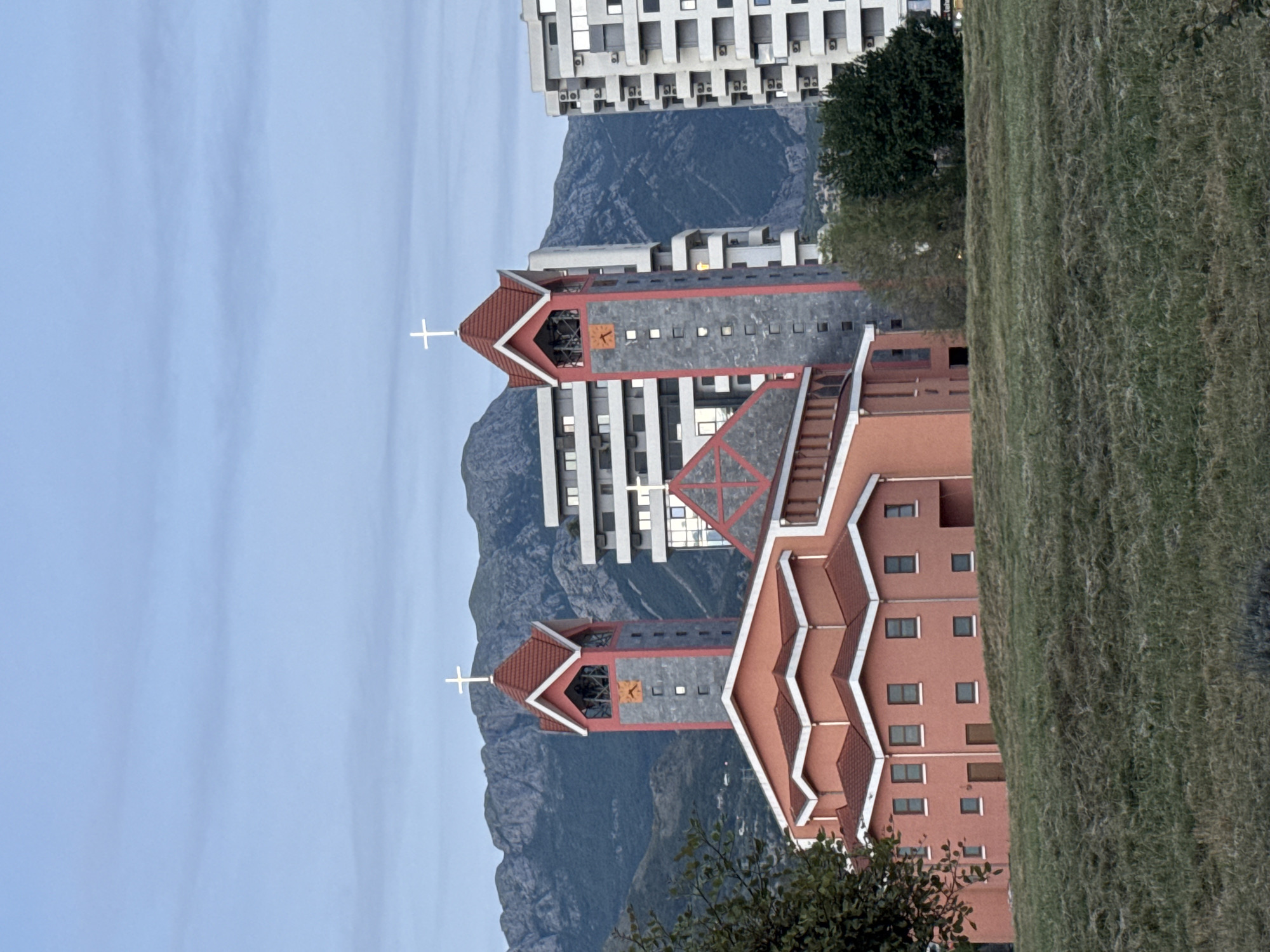 This image depicts the Cathedral of Saint Peter the Apostle (Katedrala Svetog Petra Apostola) located in Bar, Montenegro. The church, positioned centrally, features a reddish-orange facade with a complex, stepped roofline composed of red tiles and white eaves. Two prominent, symmetrical bell towers flank the main structure; these towers are constructed with gray stone-like material, bordered by reddish-orange trim, and topped with conical red roofs, each surmounted by a white Latin cross. Each tower also exhibits an orange square clock face or decorative panel and multiple small, square window-like openings. To the left of the main church structure, a lower section with a similar architectural style, featuring multiple rectangular windows and two doors, is visible.
In the background, rugged, dark green mountains rise prominently under a light blue sky with faint, horizontal cloud striations. Modern, multi-story buildings, primarily white or light gray with numerous windows, balconies, and visible air conditioning units, are situated behind and to the sides of the church, blending into the urban landscape of Bar.
The foreground consists of a gently sloping grassy area, appearing recently mown, with a mix of green and drier light brown blades. A tree branch with dark green leaves partially obscures the lower left portion of the church, and some darker green foliage is visible on the right side of the frame, behind the grassy slope. No individuals are present in the scene. The overall lighting suggests either early morning or late afternoon conditions, with soft, ambient light illuminating the subjects.