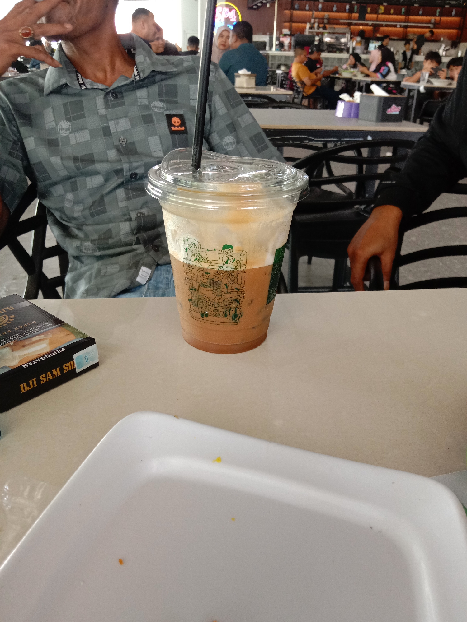 The image depicts a close-up view of a person sitting at a table in what appears to be a cafe or restaurant in Bireuen, Indonesia. The person, partially visible, is wearing a gray patterned shirt and has a ring on their finger. In the foreground, a plastic cup containing a iced coffee drink with foam is prominently featured, along with a pack of cigarettes. The background shows other patrons seated at tables and a bar area, suggesting a public establishment. The setting is indoors, with ambient lighting and a neutral color palette dominating the scene.