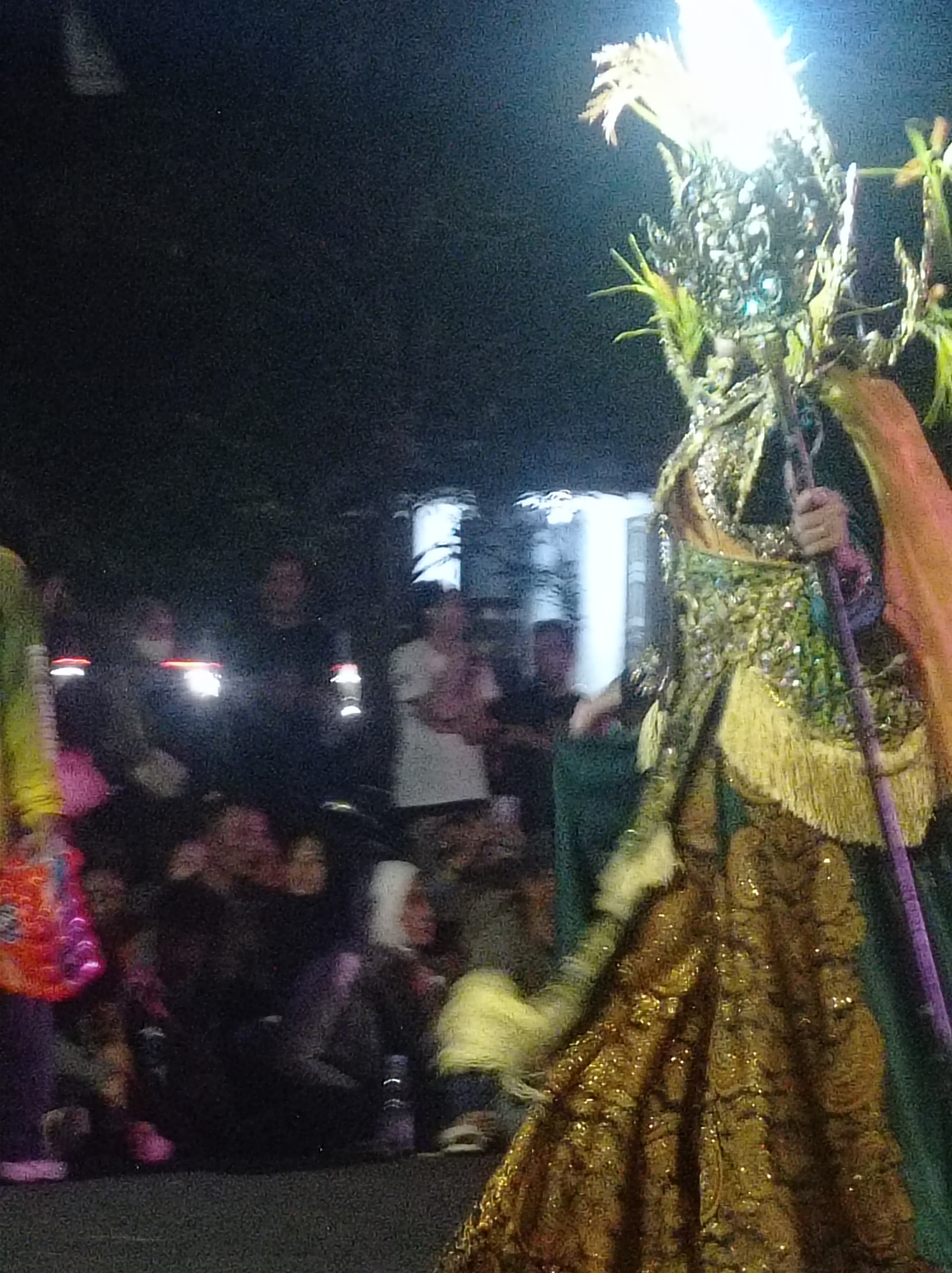 This is a nighttime, slightly blurry, medium shot of a parade or festival in Pandean, Indonesia. The central figure is a person in an elaborate, sequined costume with a tall headdress that emits a bright light. They are holding a long, purple staff. The costume is predominantly green and gold, with fringe detail. In the background, a crowd of people are visible, some holding up illuminated objects, possibly phone lights or flashlights. Buildings with glowing windows can be seen in the far background. The overall impression is of a lively, illuminated event taking place after dark.