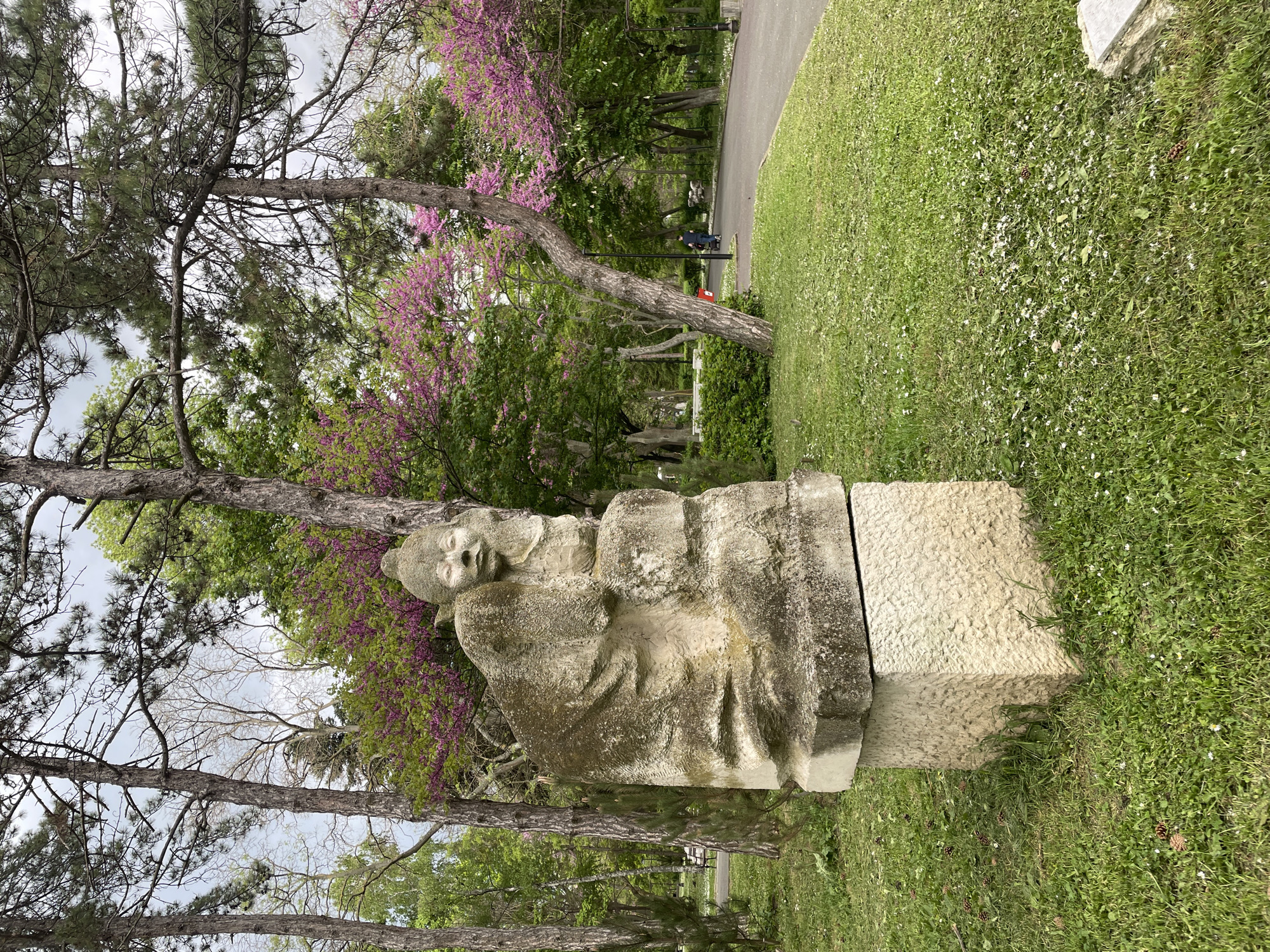 The image shows a stone sculpture in a park setting. The sculpture appears to depict a human figure, though weathered and partially obscured. The sculpture is situated on a stone pedestal. The environment includes trees, some with purple blossoms, and a grassy area. A paved walkway is visible in the background with a person. The sculpture is located in Burgas, Bulgaria.