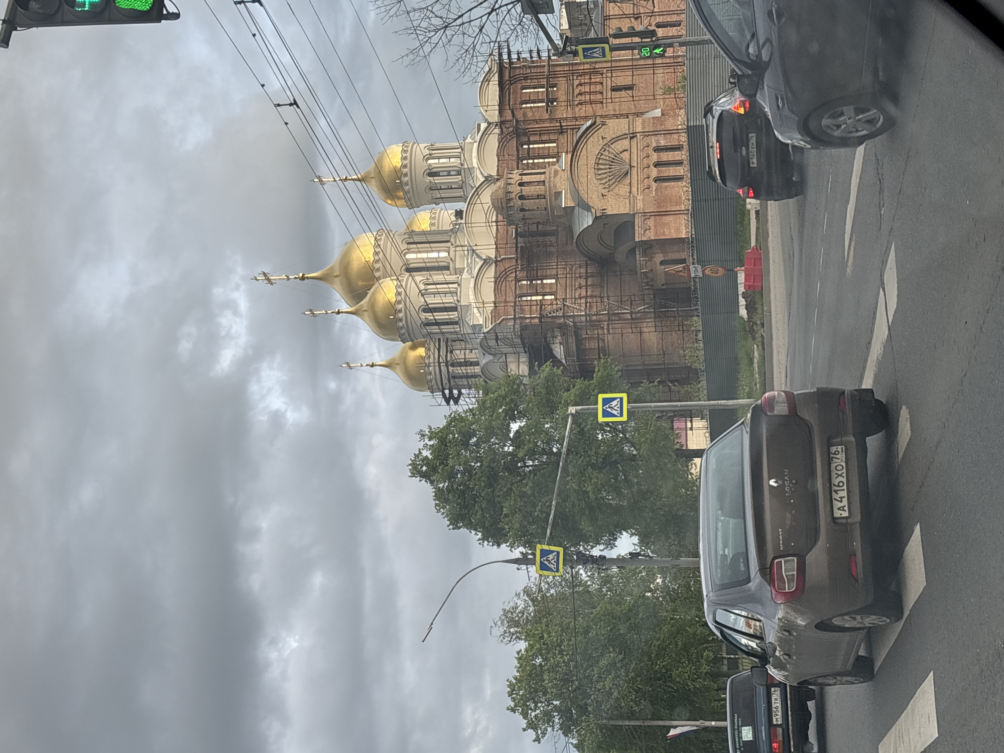 The image shows a street scene with a building in the background. The building is a cathedral with multiple golden domes and is under construction or renovation, as scaffolding is visible. A green traffic light is visible in the upper left corner of the image. Several cars are on the street, including a brown Renault Logan with the license plate A416XO76. A pedestrian crossing sign is visible near the center of the image. The sky is overcast with dark clouds. The city is Rusanovo, Russia.