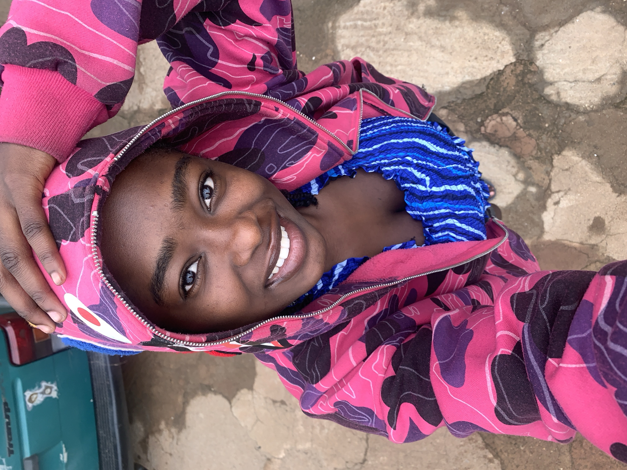 A young woman with a dark complexion smiles directly at the camera in what appears to be a selfie, with her left hand (from her perspective) resting on the top of her head, holding up the hood of her jacket. She has dark eyes and is smiling broadly, revealing her teeth. She is wearing a pink and purple camouflage-patterned zip-up hoodie featuring abstract shapes and a distinct white, red, and black shark-face graphic on the right side of the hood (viewer's left). A silver zipper runs down the front of the partially open hoodie. Underneath, a blue and light blue horizontally striped top is visible. The setting is outdoors, in the city of Rafin Par, Nigeria. The ground beneath her is an uneven, cracked surface composed of light brown concrete or packed earth. In the upper left background, part of a faded teal or dark green vehicle, possibly a pick-up truck or SUV, is visible, showing wear and bearing the partial inscription "Tranup" on its rear.