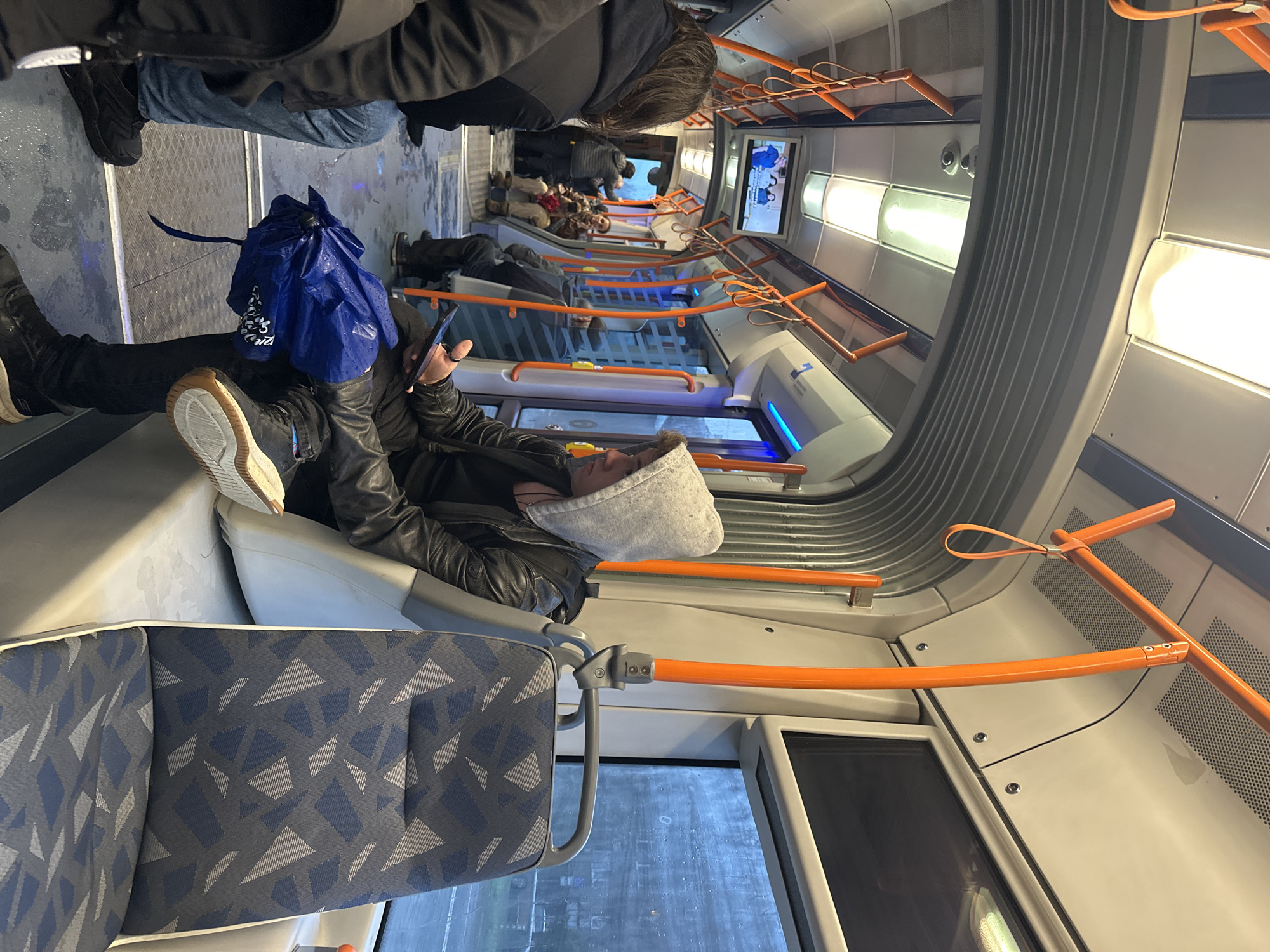 Inside a public transport vehicle, possibly a bus or train, in Sofia, Bulgaria. A person is lying down on a row of seats. They are wearing a black leather jacket, dark pants, and shoes with light-colored soles. They have a grey hood over their head and are looking at a smartphone in their hands. A blue bag is resting on their lap. Other passengers are visible in the background. The vehicle has rows of seats upholstered with a blue and grey geometric pattern. Orange handrails are positioned along the upper part of the vehicle. Windows are visible along the side, and a screen displays content towards the front. The interior is lit with ceiling-mounted lights.
