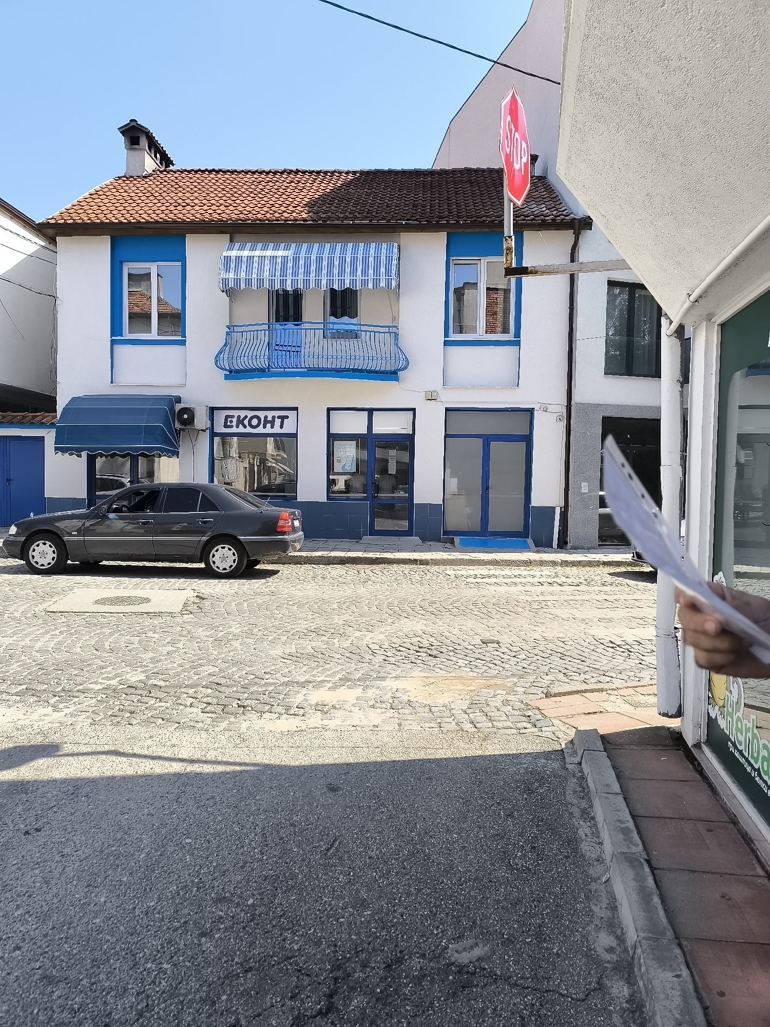 The image shows a street scene in the city of Gotse Delchev, Bulgaria. In the foreground, a gray sedan is parked on a cobblestone street. Behind the car, a white building with blue trim and awnings stands. A sign with the letters "EKOHT" is visible on the building. To the right, a stop sign is posted on a pole. The sky is clear and blue. The image appears to be taken on a sunny day.