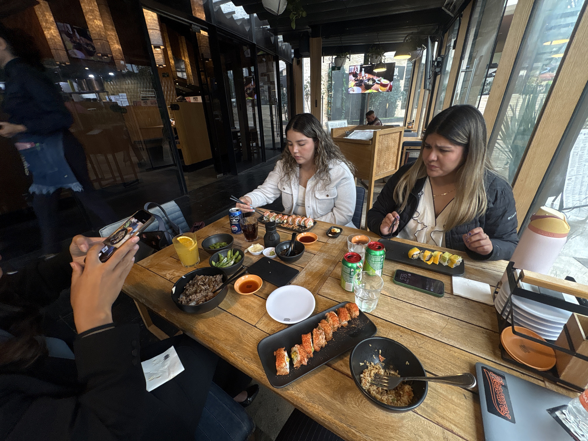 The image depicts a dining scene at a restaurant in Mexico City, Mexico. Two women are seated at a wooden table laden with sushi, rice dishes, edamame, and drinks. A third person is taking a photo of the food with their phone. Other objects on the table include condiments, plates, glasses, and cutlery. A laptop is visible at the table's edge. The restaurant has large windows offering natural light and views. A TV screen showing a broadcast is visible in the background. Other diners are present, as well as a restaurant server. The atmosphere appears casual and contemporary.