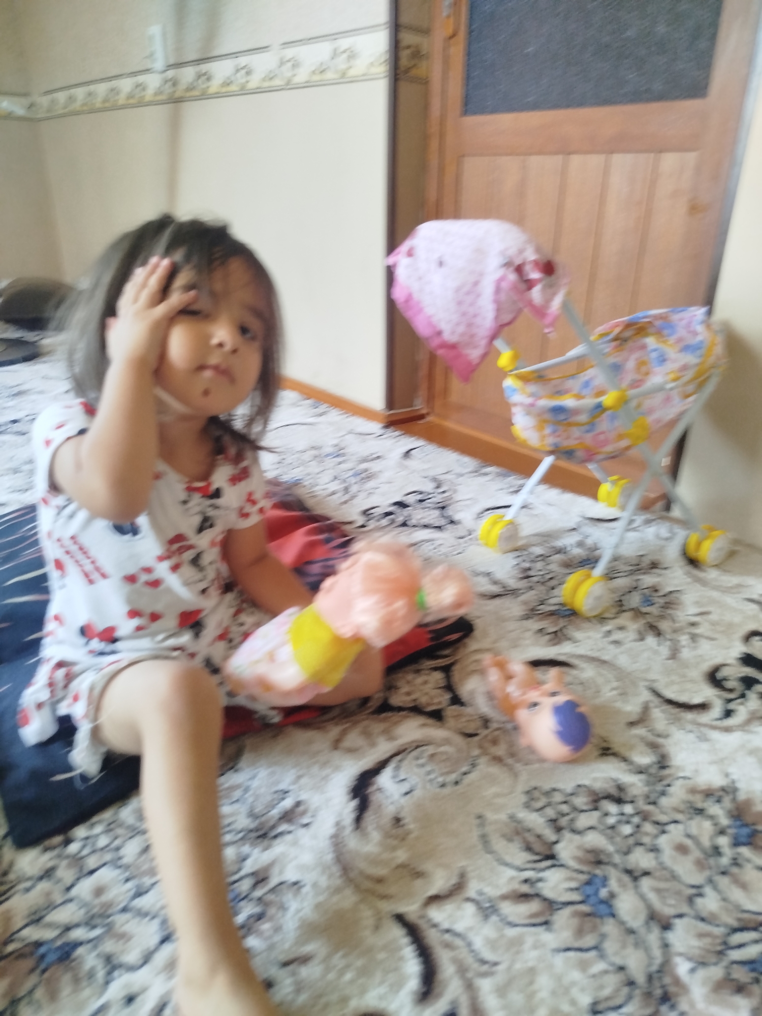 A young child with dark hair is sitting on a patterned rug in a room, with their hand covering part of their face. They are wearing a white t-shirt with a red and black pattern. In front of the child is a toy baby carriage and two baby dolls. The room appears to be a living room or bedroom, with a wooden door in the background and a patterned wallpaper on the walls. The scene suggests a domestic setting, possibly in the city of Kokand, Uzbekistan.