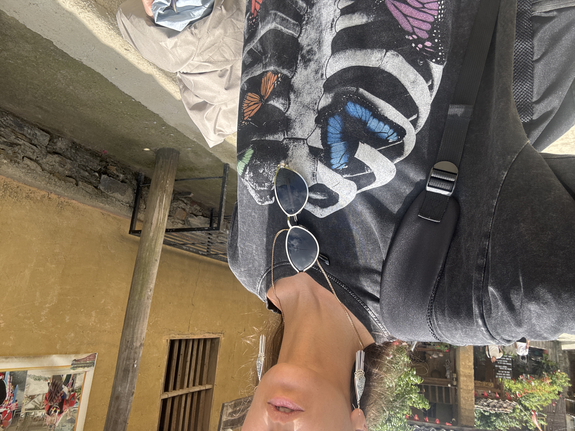 The image shows a person wearing a grey t-shirt with a skeleton print adorned with butterflies, along with silver earrings and sunglasses hanging from a gold chain. The person is wearing a black backpack.
The background includes a yellow wall with a window, a wooden post, and a framed picture. The scene appears to be outdoors, possibly in a courtyard with greenery. The presence of Vietnamese architecture and vegetation suggests the city of Lũng Cú Commune, Vietnam, as the location. A glimpse of people and buildings in the far background further supports an outdoor setting in a populated area.