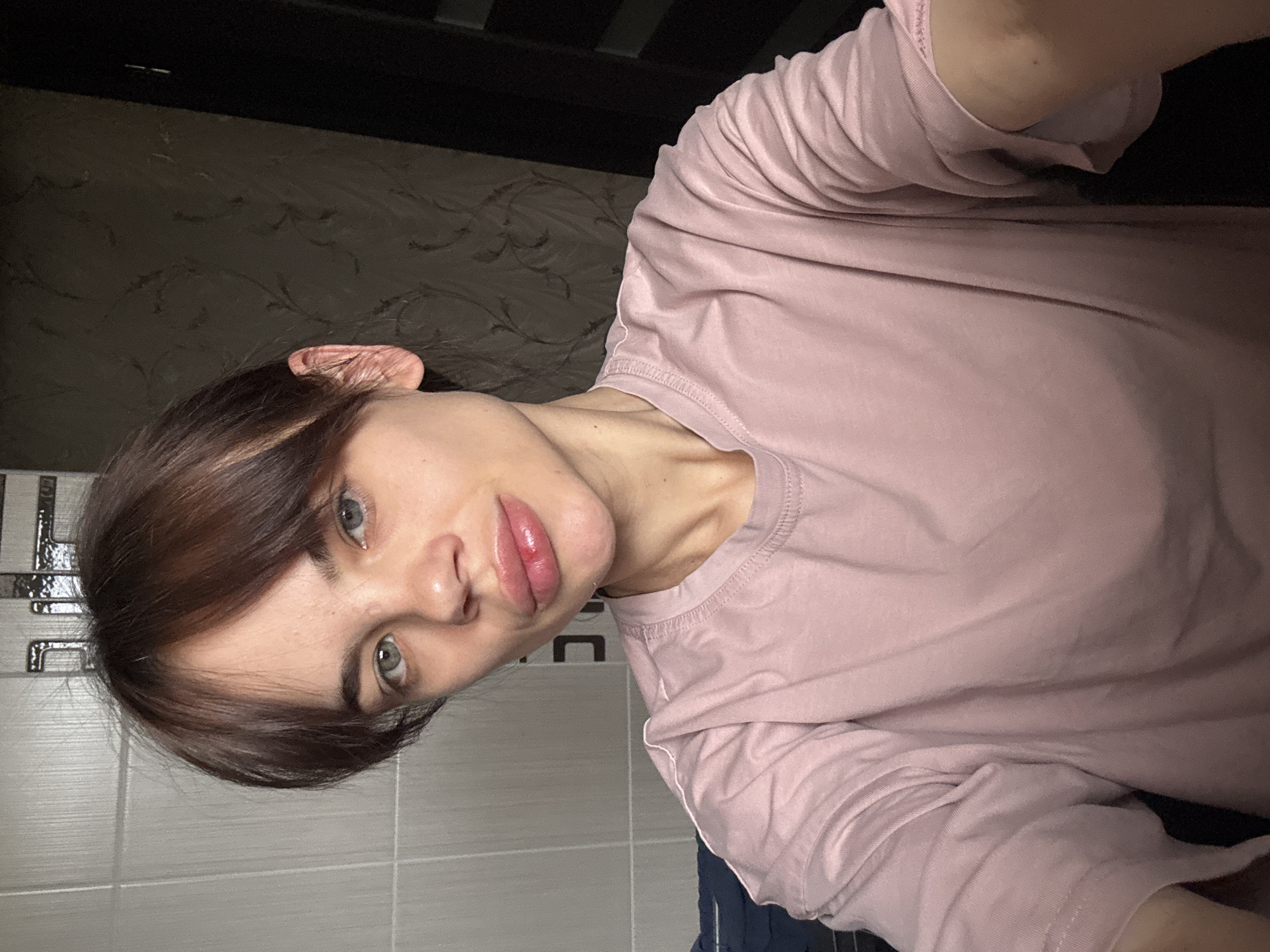 A woman with brown hair and gray eyes, wearing a pink t-shirt, is looking at the camera. The background is a wall with dark wallpaper and a tiled wall with a metallic trim. The location information is not discernible from the image.