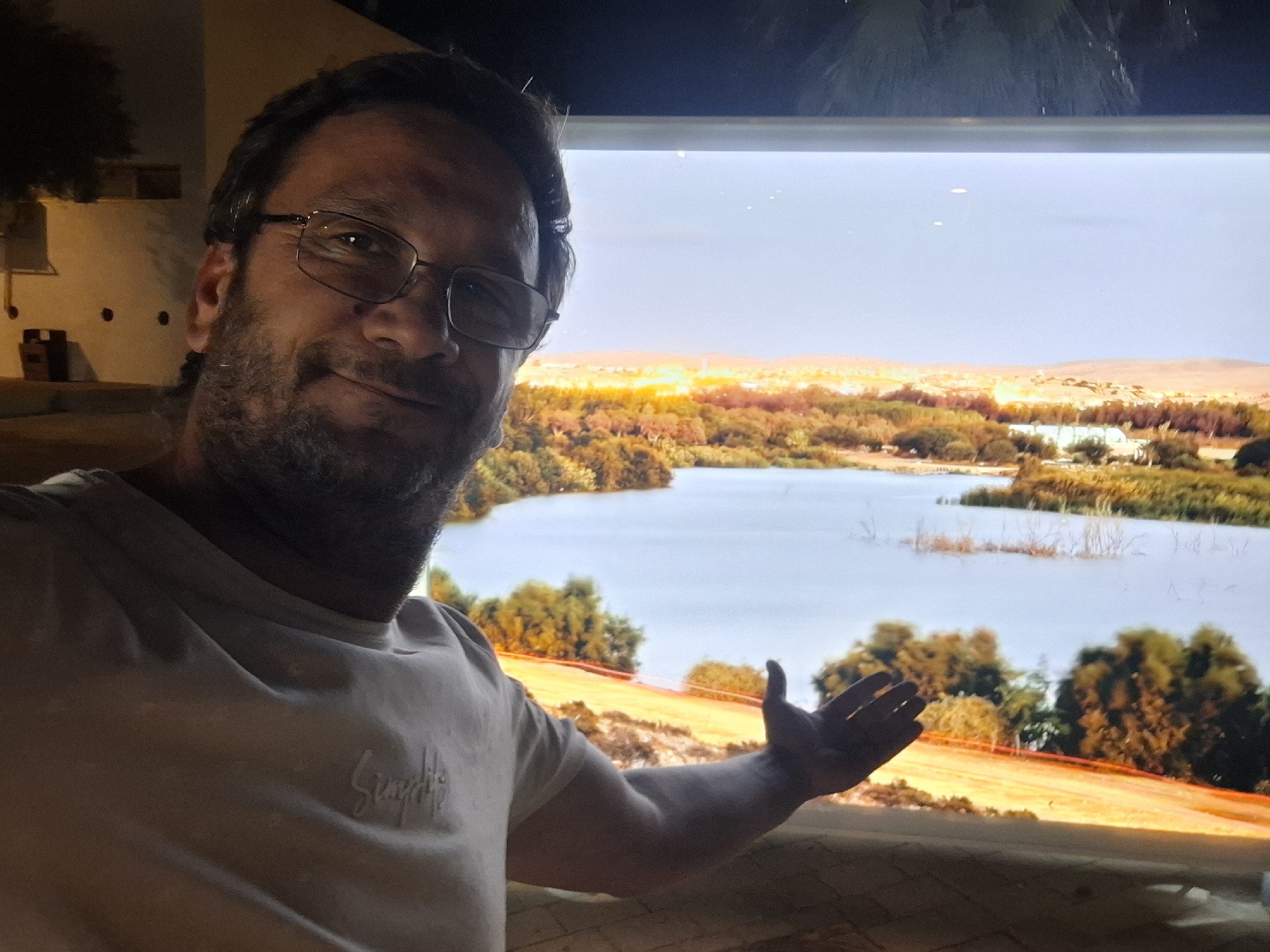 A man with glasses and a beard takes a selfie with a large screen displaying a landscape behind him. The screen shows a body of water with trees and buildings in the background under a clear sky. The man is wearing a light-colored t-shirt with an embroidered logo on the left chest. He is smiling and gesturing with his right hand. The image appears to be taken in the evening, with artificial lighting visible in the background, possibly indicating an indoor location in Yerocham, Israel, perhaps a public space or a private residence with a large display.