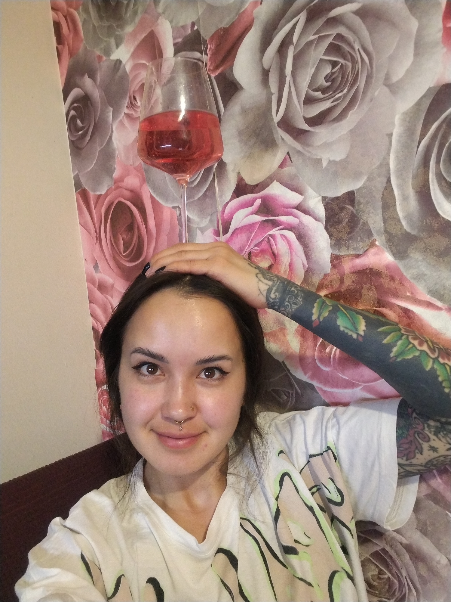 A woman is posing for a selfie indoors. She has brown hair, a nose piercing, and is wearing a white t-shirt with a light-colored abstract design. Her right arm is raised above her head, and she has a full-sleeve tattoo on that arm. A wine glass filled with a red liquid is balanced on top of her head. The background is a wall covered in a floral wallpaper pattern. The location in the city of Noviy, Russia cannot be determined from the image.