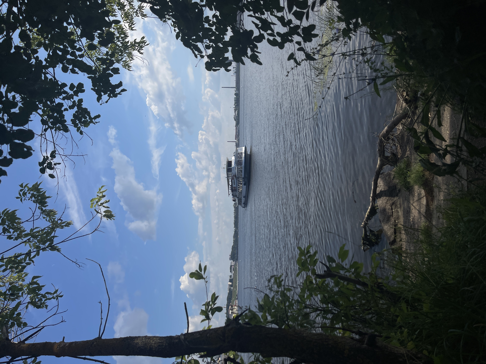 A ferry is seen on the Izh River in Izhevsk, Russia. The sky is partly cloudy and the water is calm. There are trees in the foreground, and buildings in the background. The ferry is white with a red stripe and has a capacity of about 50 people. The ferry is moving from left to right across the image.