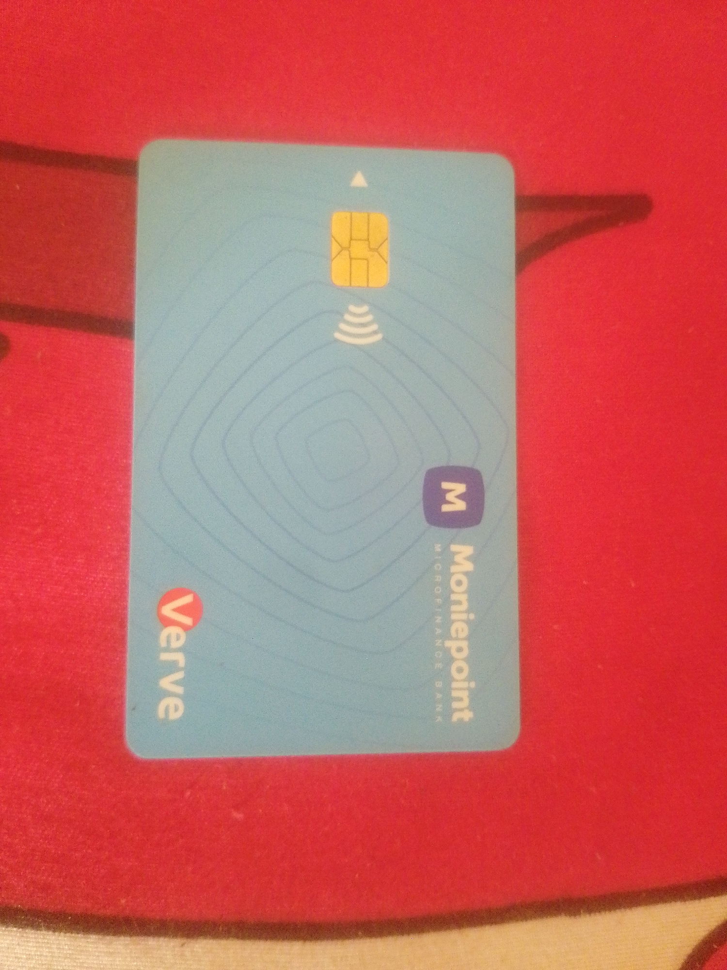 The image shows a light blue debit card with a golden chip and a contactless payment symbol. The card has the "Verve" logo on the bottom left and the "Moniepoint" logo and name on the right. The card is lying on a red textured surface. The scene does not provide any information or context about the city of Danbazau, Nigeria.