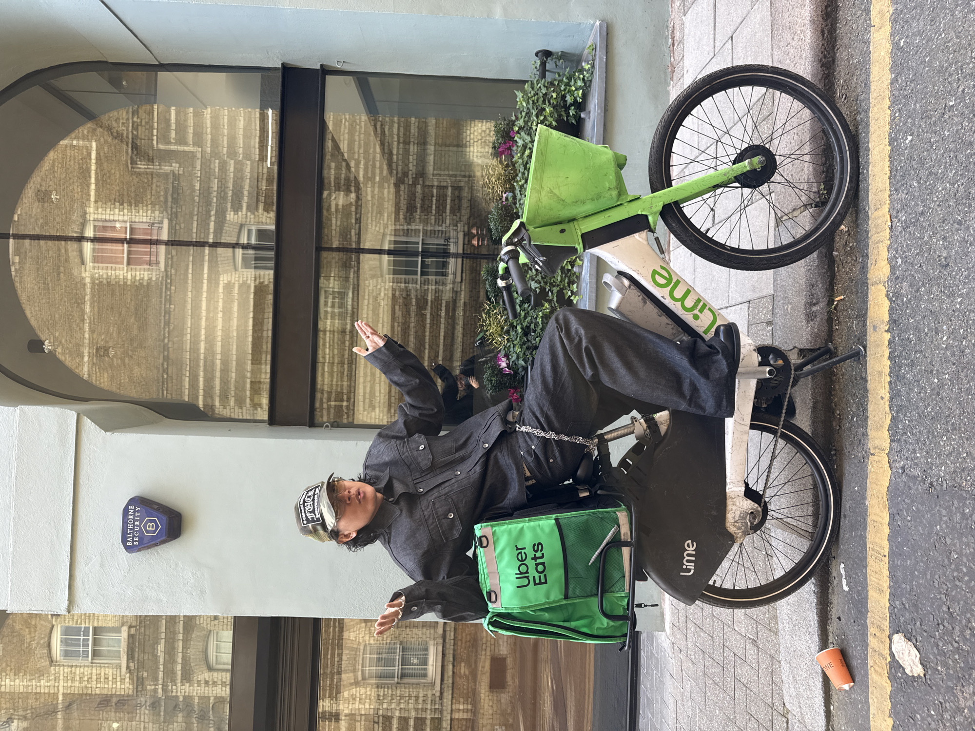 An individual is casually posing on a green and white Lime e-bike on a paved sidewalk next to an asphalt road, in what appears to be an urban setting, likely London, United Kingdom. The person is dressed in a dark denim jacket, dark loose-fitting trousers, a cap, and sunglasses. They are looking towards the camera with both hands raised, palms open, suggesting a playful or confident gesture. A prominent green Uber Eats delivery bag rests on the back rack of the e-bike.
The background features a light-colored building wall with a large arched window reflecting the facade of an older brick building across the street. Beneath the window, there are potted plants with some purple flowers. To the left of the person, a blue hexagonal security alarm box is mounted on the wall, displaying the text "BALTHORNE SECURITY" and a "B" logo. The cap worn by the individual also has visible text, including the word "FUCK" among other partially obscured words.
The e-bike itself is branded with the "Lime" logo on its frame and front mudguard. The road beside the sidewalk has a distinct yellow line, characteristic of UK road markings. On the ground near the e-bike's front wheel, there's a discarded orange cup with faint text, possibly "ONE", and some litter. The scene is brightly lit by natural daylight, indicating it is daytime.