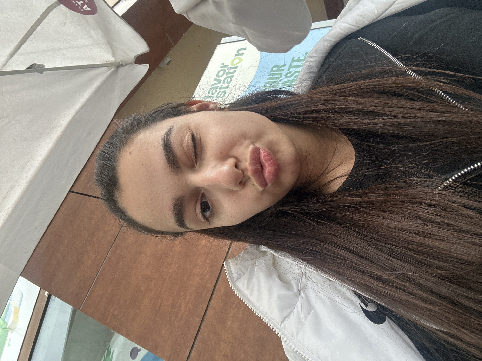 A young woman is depicted in a close-up selfie, winking with her left eye (viewer's left) and making a playful "duck face" expression with puckered lips. She has long, straight dark brown hair that falls across her shoulders and prominent dark eyebrows. She is wearing a black top or jacket with a silver zipper, layered with a white puffer jacket or vest, which shows a partial Nike swoosh logo. A slender silver necklace is visible around her neck.
The setting appears to be an outdoor or semi-outdoor cafe or seating area in Plovdiv, Bulgaria. A large white umbrella provides shade in the upper left of the frame. Behind her, there are brown wooden panels, likely part of a building's exterior. Further in the background, through a glass partition or on a sign, partial text is visible: "Flavor Station" (specifically "lavor Station") in green, and "YOUR TASTE" (partially "YOUR ASTE") in green and blue lettering respectively. Upside-down text, possibly "AT", is also visible on the white umbrella fabric. The natural light suggests it is daytime. Her expression conveys a sense of fun and playfulness.
