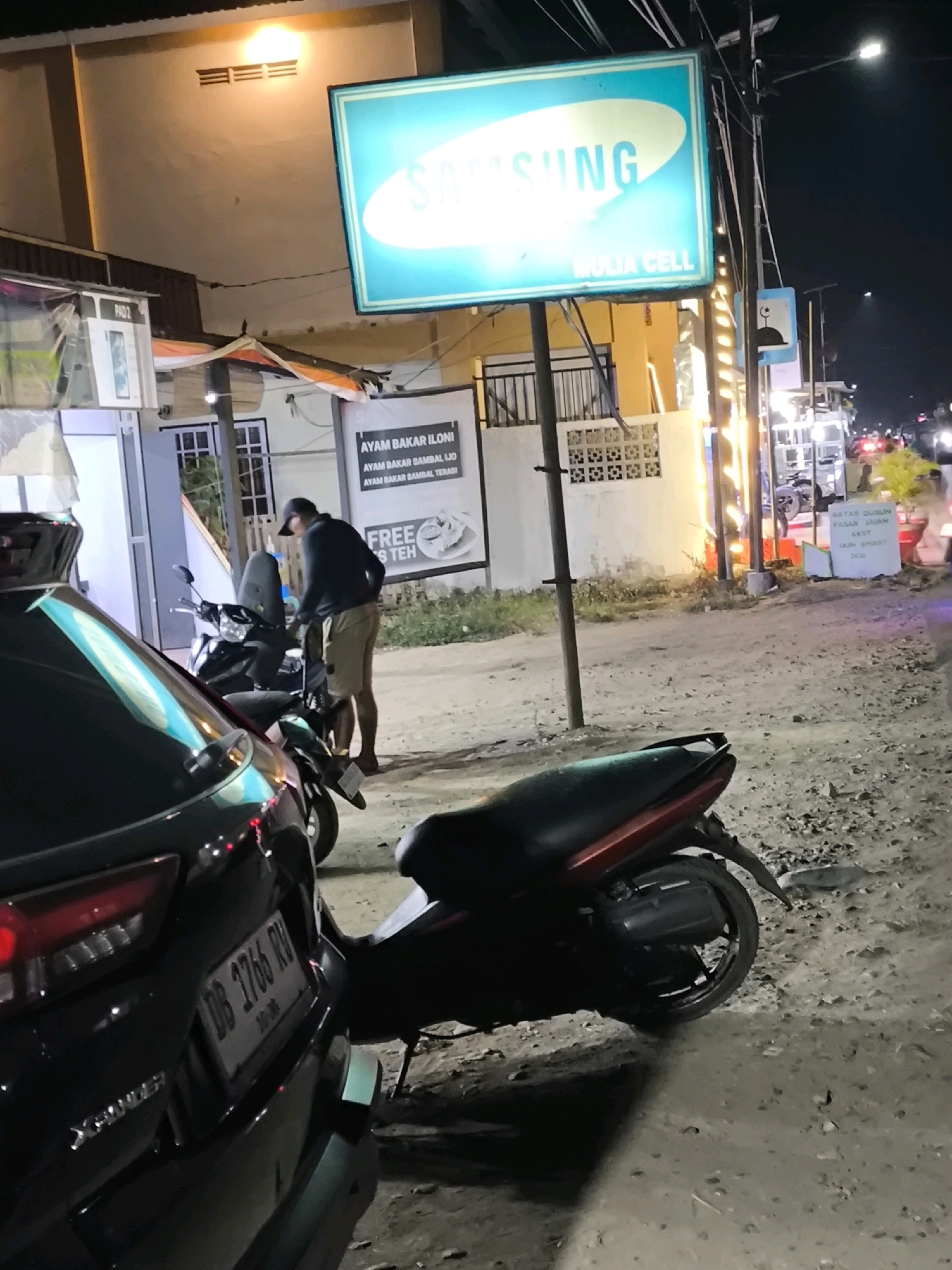 An exterior shot captures a street scene at night in Moluo, Indonesia. A person wearing a dark long-sleeved shirt and khaki shorts, a black baseball cap, and dark shoes stands next to a black motorcycle. The motorcycle has a red and black seat and a gray rear fender. To the left of the motorcycle, the rear of a black car is visible, displaying a license plate that reads "DB 1766 RV". Behind the car and motorcycle, a building with a bright orange wall is partially visible, along with a large neon sign that says "SAMSUNG" in white text on a turquoise background, and below that, "MULIA CELL". Further back, another sign reads "AYAM BAKAR ILONI AYAM BAKAR SAMBAL IJO AYAM BAKAR SAMBAL TERASI" and "FREE ES TEH". A streetlight with a mosque symbol is visible on a pole. The ground is unpaved and covered with gravel. The overall lighting suggests it is nighttime with artificial light sources illuminating the area.