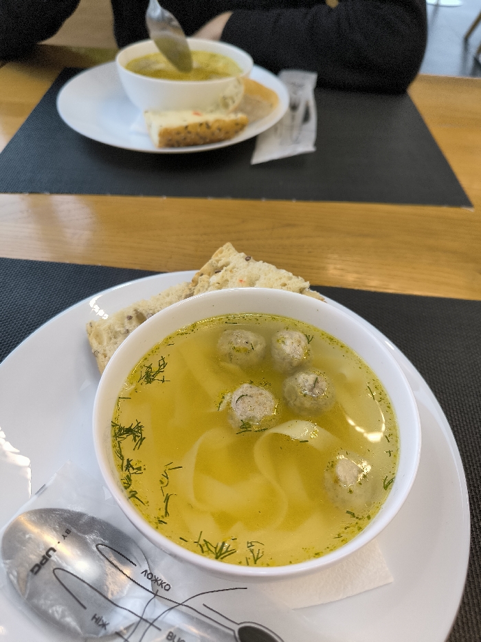 Two bowls of soup are on a table with a wooden section and a dark mat. The bowl in the foreground contains chicken noodle soup with small meatballs and fresh dill. A slice of bread and a spoon in a plastic wrapper lie beside the bowl. The background bowl holds a light green soup, accompanied by a slice of bread. The scene suggests a restaurant setting, featuring two different soup options.