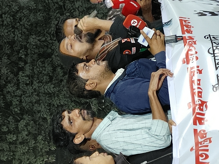 The image depicts an outdoor night-time gathering in Bangladesh, likely a press conference or public address, set against a backdrop of dense green foliage. Several adult males are visible from the chest up, positioned behind a white banner with red Bengali text. A central figure, bearded and wearing a dark hooded garment with a striped scarf, is actively speaking into a black handheld microphone, his eyes closed and mouth open in mid-speech. To his immediate left, a bearded man in a dark blue jacket and white shirt observes to the right. Further left, a thin man with a goatee and dark, curly hair, wearing a light blue checkered shirt, also looks to the right. Partial views of other individuals are visible on both sides. Multiple media microphones, including those with logos for RTV, DBC News, and Desh TV, are directed towards the speaker, held by unseen individuals. The banner in the lower foreground displays Bengali text, partially visible as "આતંક રાષ્ટ્રવ્યાપી ઉજ્જડ કરોન!" (Atanka rastrabayapi ujjar karoon!), translating to "Destroy nationwide terror!" or "Stop state terrorism!". A specific location within the city of Dhaka is not discernible from the visual evidence.