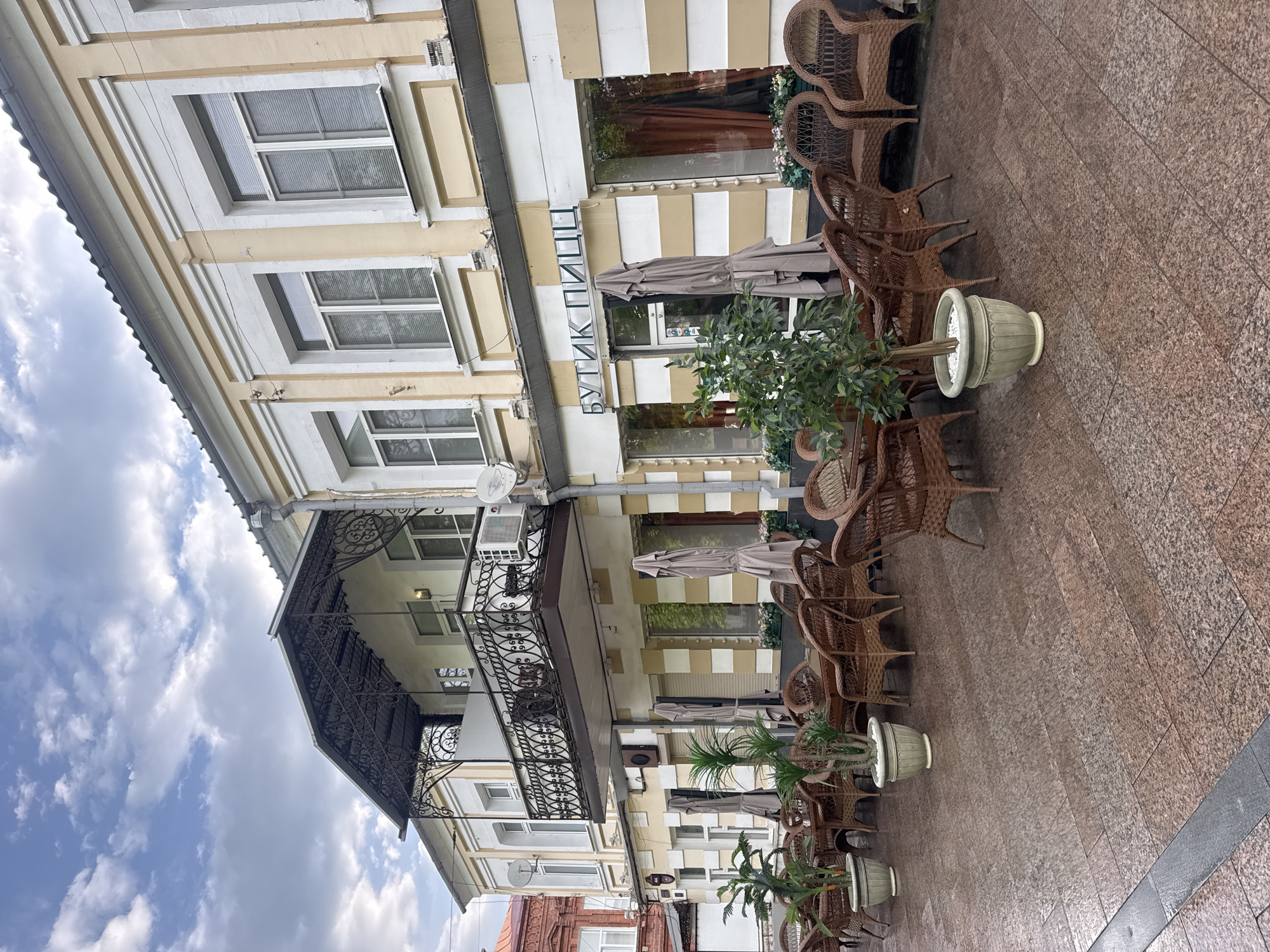 The image depicts a street view in Vladikavkaz, Russia, featuring a two-story building with a commercial establishment on the ground floor. The building facade is painted in alternating stripes of yellow and white, with multiple windows and a balcony adorned with decorative wrought iron. A sign in Russian, vertically oriented, reads "БУТИК ПИЦЦ" (Boutique Pizza).
Several brown wicker chairs are arranged outside the establishment, interspersed with potted plants of varying sizes and species. Partially visible umbrellas suggest an outdoor dining area.
The street is paved with reddish-brown tiles. The sky is visible in the upper left corner, showing a partly cloudy day. The overall impression is of a European-style street cafe. No people are visible in the image.