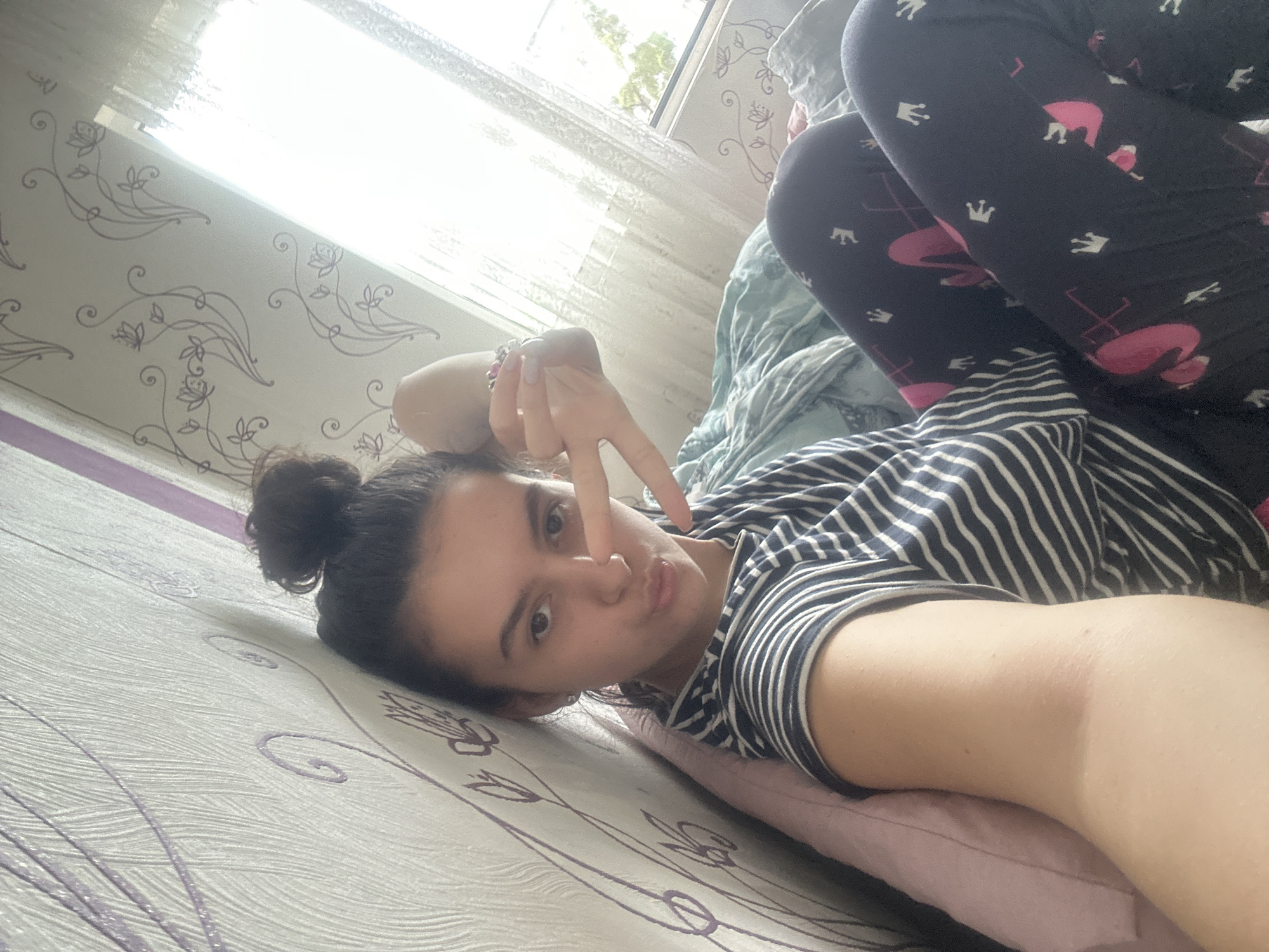 A person is lying on a bed in a room with floral-patterned wallpaper. The person is wearing a striped top and leggings with flamingo and crown patterns. They are making a peace sign with their hand. The location is Unknown, Unknown. A window is visible in the background.
