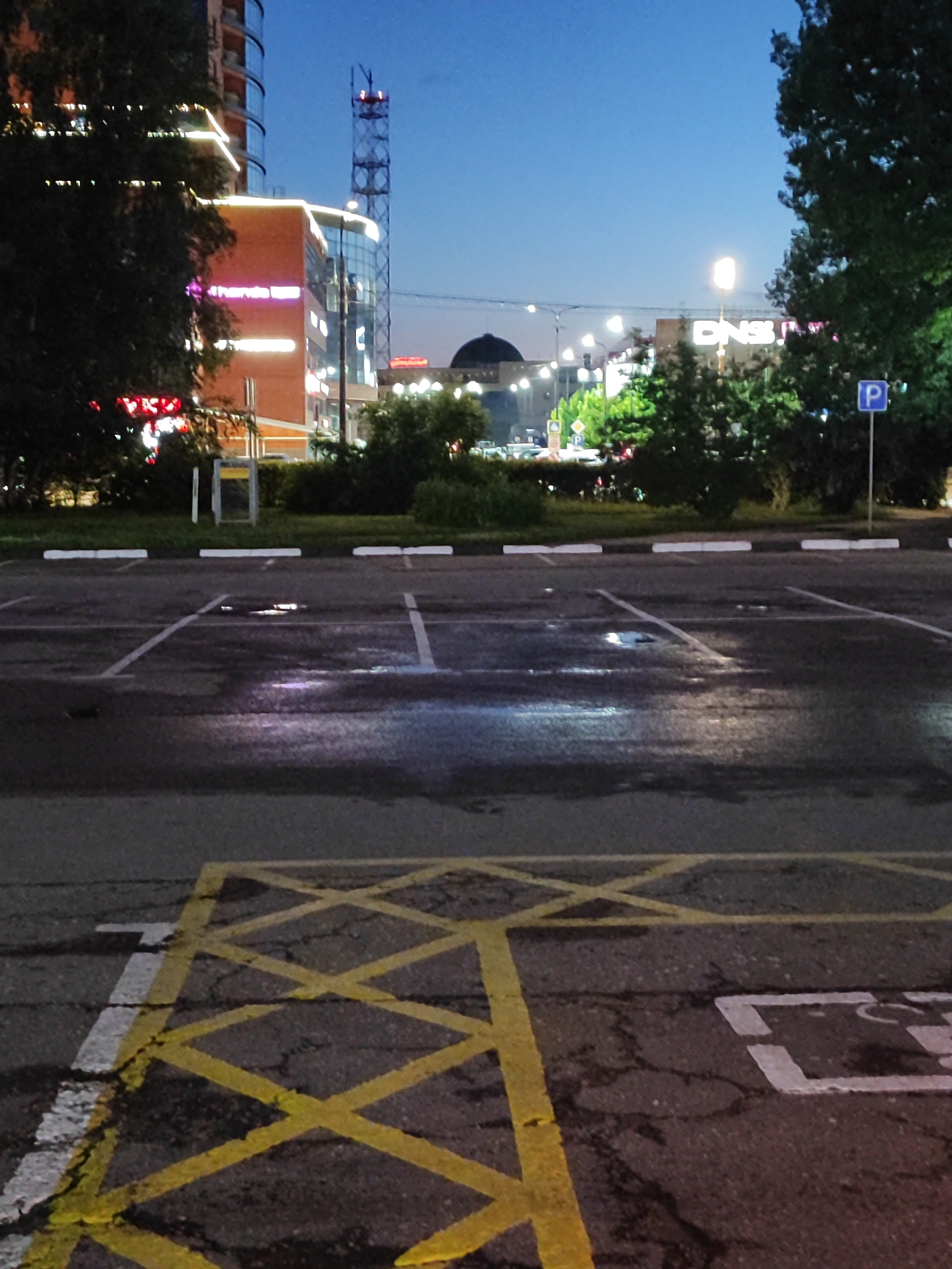 This is a medium-angle, eye-level shot of an outdoor parking lot at dusk, with a city skyline in the background. The parking lot asphalt is wet and reflects the ambient light. Yellow criss-cross markings and white lines indicate parking spaces. Some white parking bollards are visible. In the background, there is a modern building with illuminated signage and lights along its facade, a tall radio tower, and a domed structure. Trees are present on the right and left sides of the image. A blue parking sign with a white "P" is visible on the right. The sky is a deep blue, transitioning into twilight.